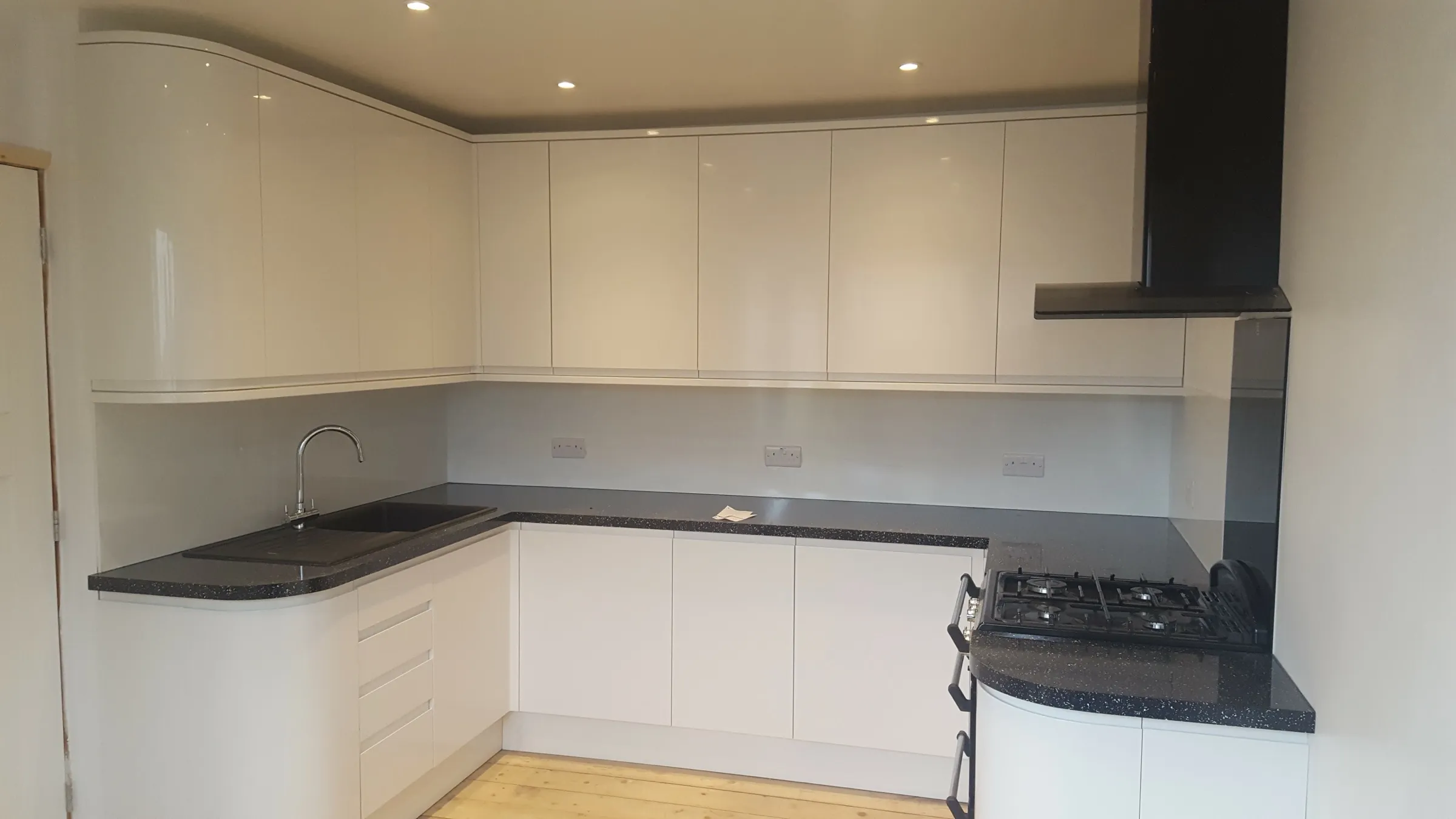 L-shaped white gloss kitchen with curved corner units, dark speckled worktops and a black chimney hood over a gas cooker
