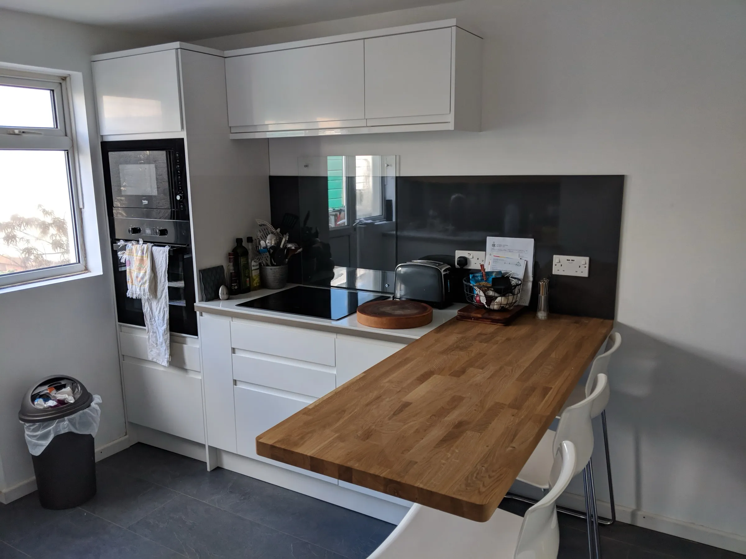 White handleless kitchen with smoked glass splashback, built-in black oven and microwave, induction hob and a solid oak breakfast bar with white stools