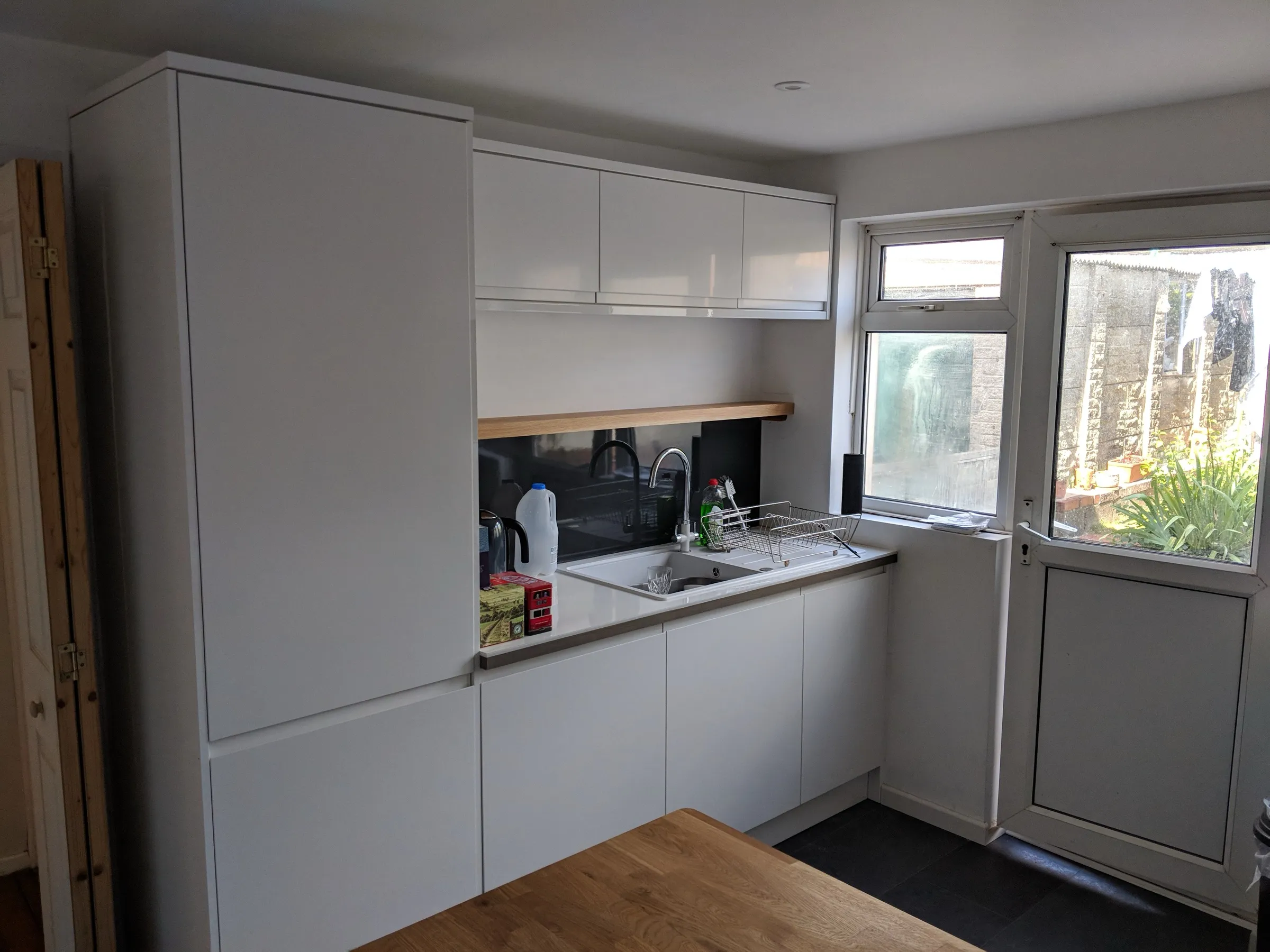 Tall white units with integrated appliances, white sink beneath a floating oak shelf and smoked glass splashback, next to a part-glazed door to the garden