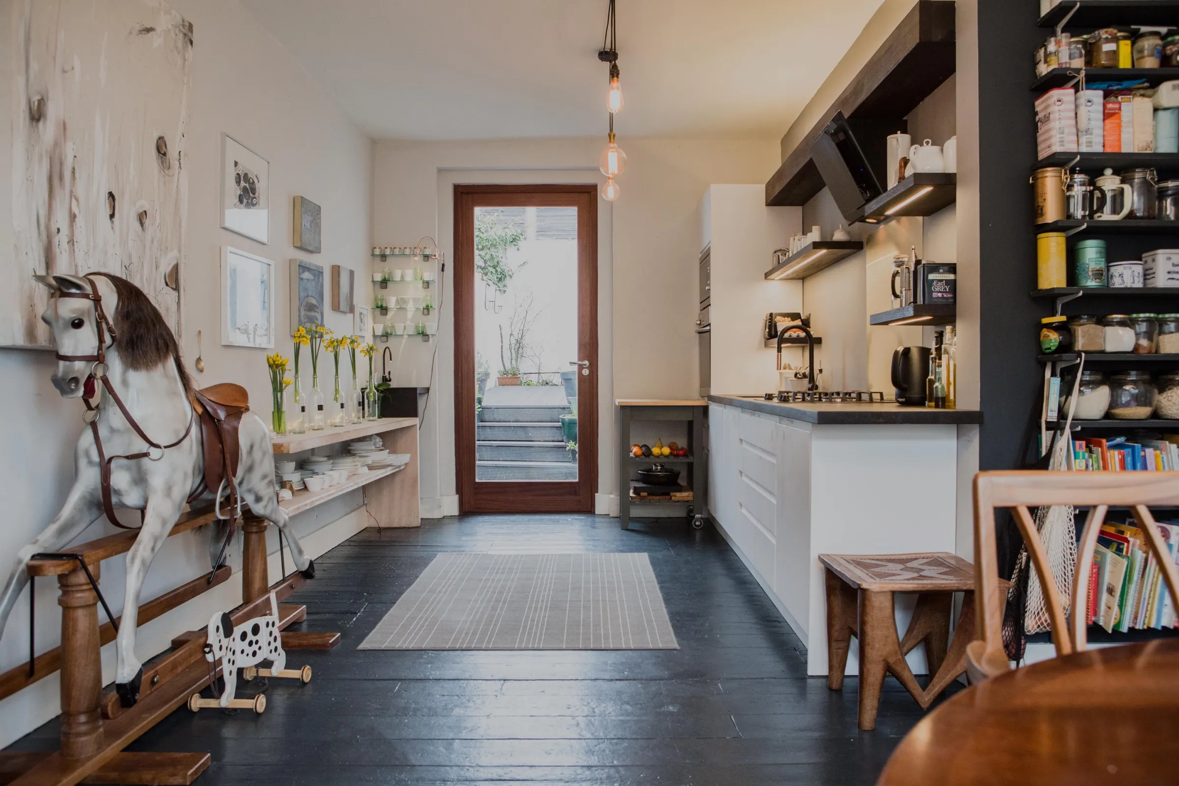 Galley view of the kitchen towards the glazed garden door, with white handleless units and dark floating shelves on one side and a gallery wall with an antique rocking horse opposite