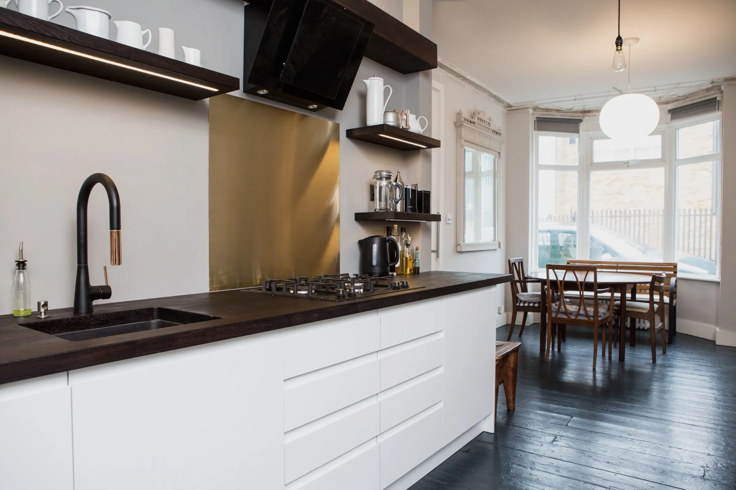 White handleless units beneath a dark-stained solid timber worktop, with matt black tap, brushed brass splashback and black angled extractor, looking through to the dining table in the bay window