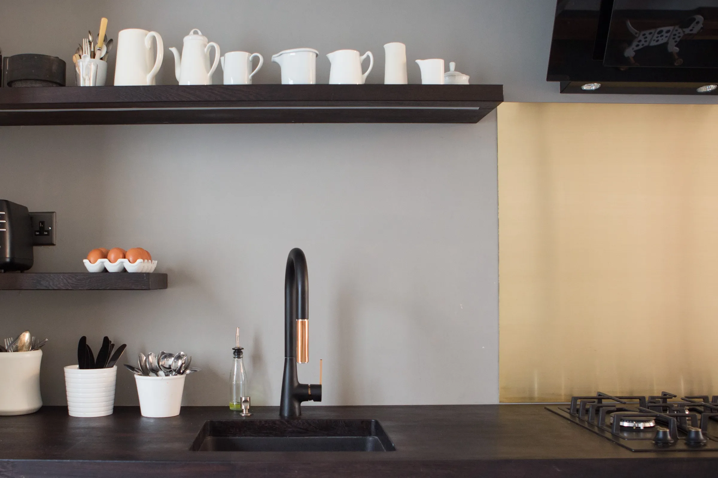 Detail of the undermounted sink and matt black tap with copper trim, below a dark floating shelf of white jugs with integrated lighting and the brass splashback beside the hob