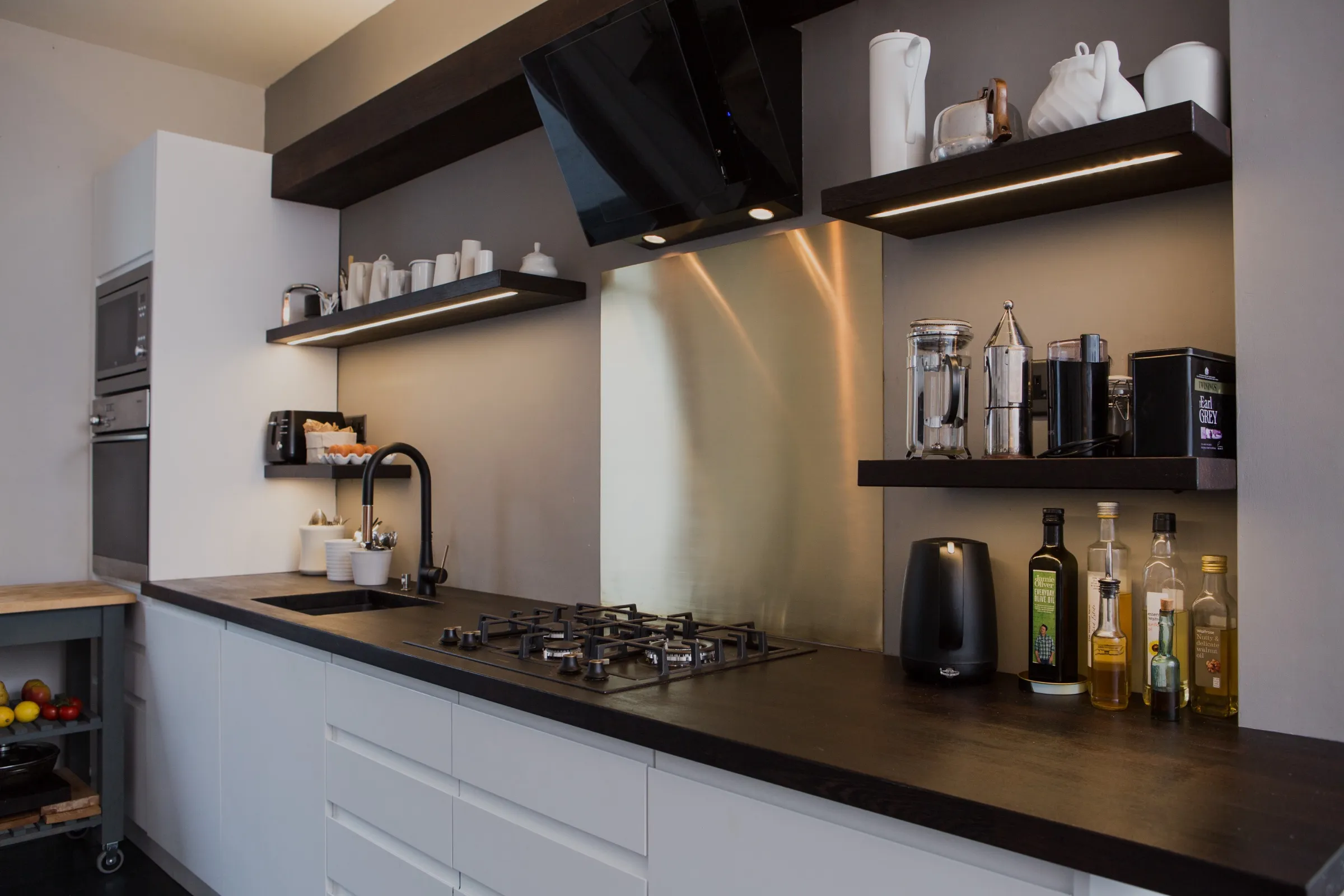 The full kitchen run from the dining end, showing built-in ovens in a tall white unit, a five-burner gas hob in the dark timber worktop, brass splashback and staggered floating shelves