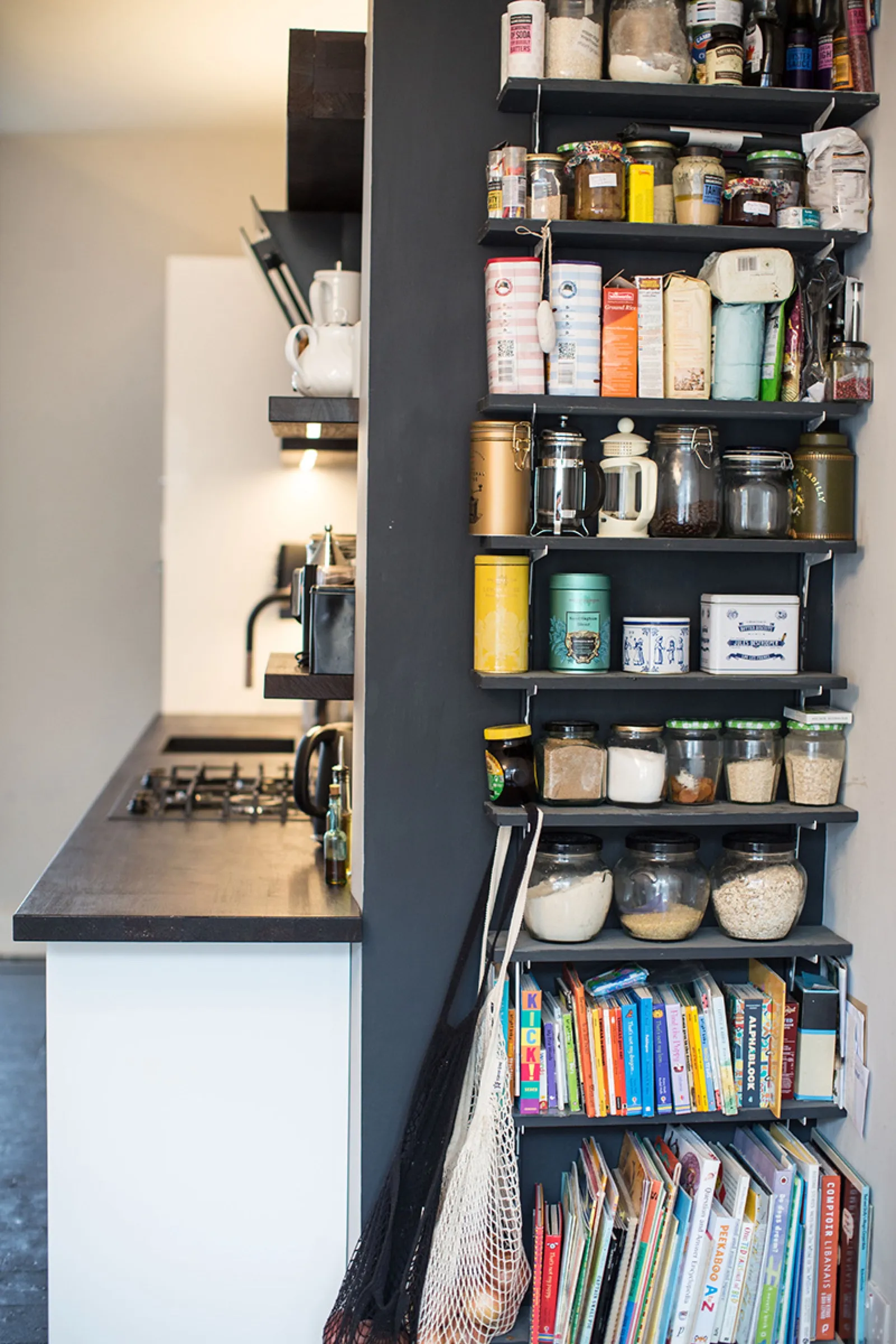 Open pantry shelving on a dark feature wall beside the kitchen, stacked with jars and cookbooks