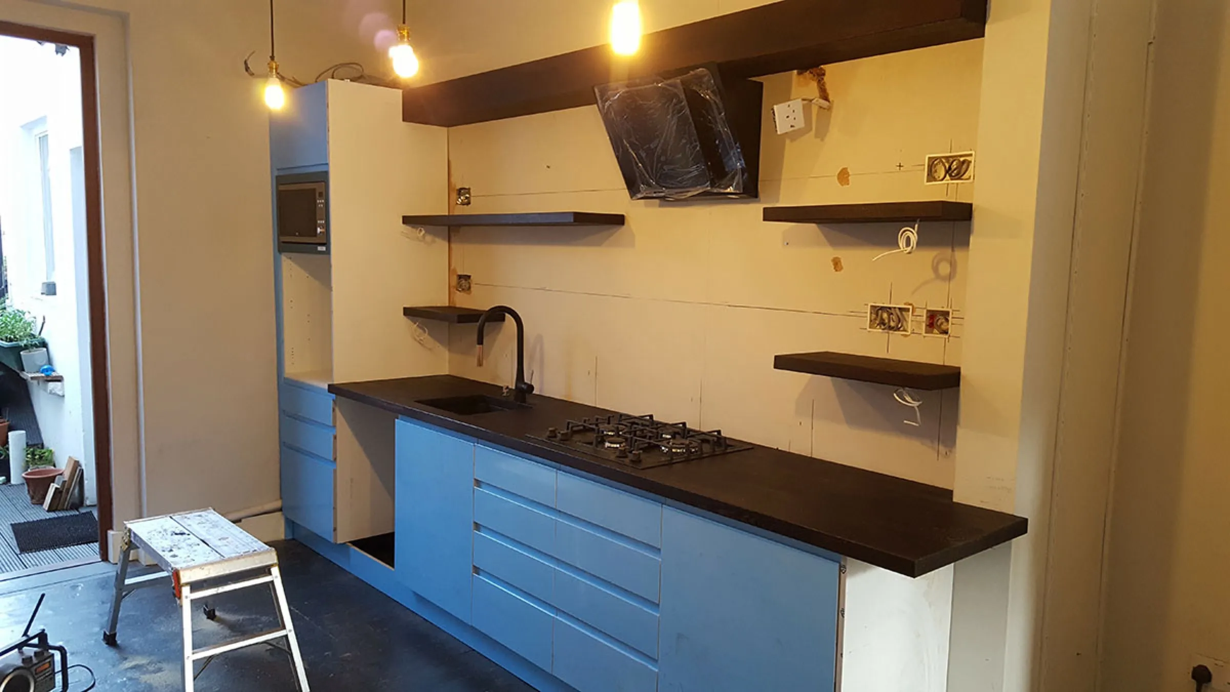 The same kitchen during installation: units in protective film, dark timber worktop and floating shelves being fitted