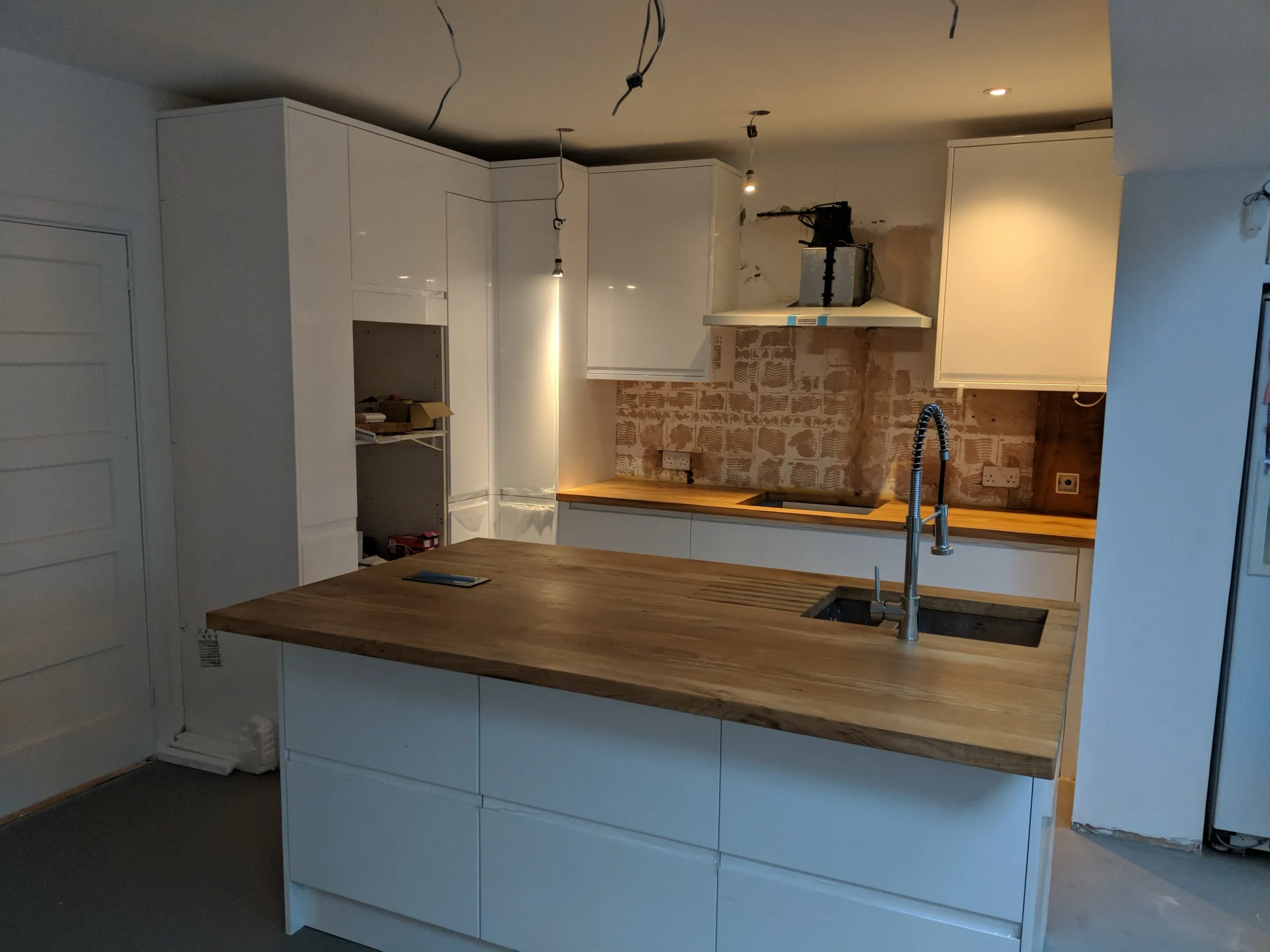 Open-plan kitchen mid-installation with white gloss handleless units, timber-topped island with undermounted sink and pull-down spray tap, and chimney extractor hood