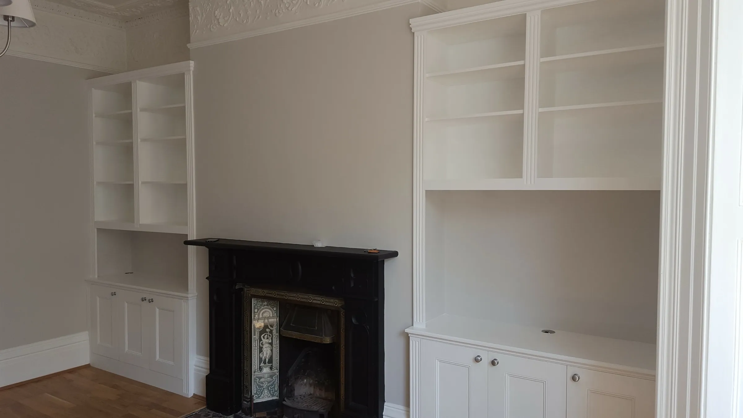 Classic white alcove dressers with panelled cupboards and open shelving either side of a black Victorian fireplace beneath ornate cornicing