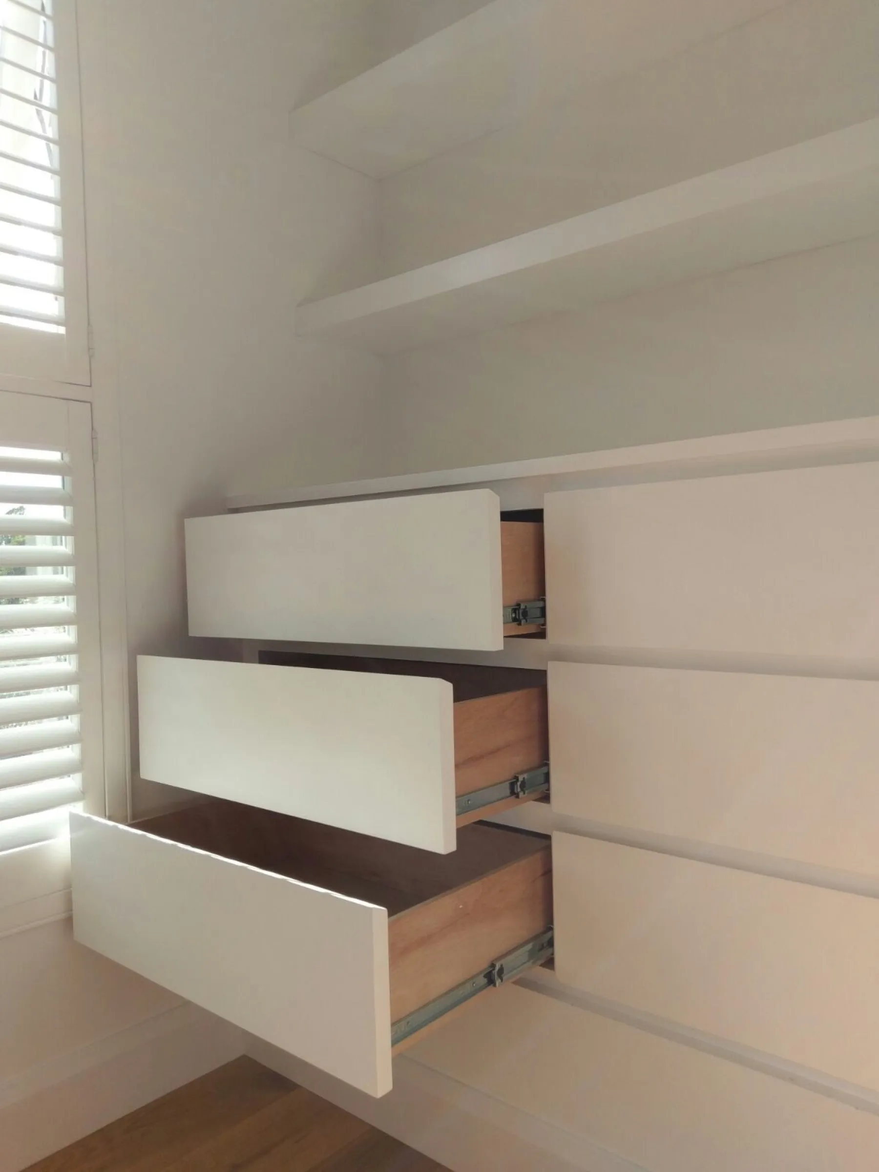 Fitted chest of drawers with plywood drawer boxes shown open, beside a window with plantation shutters