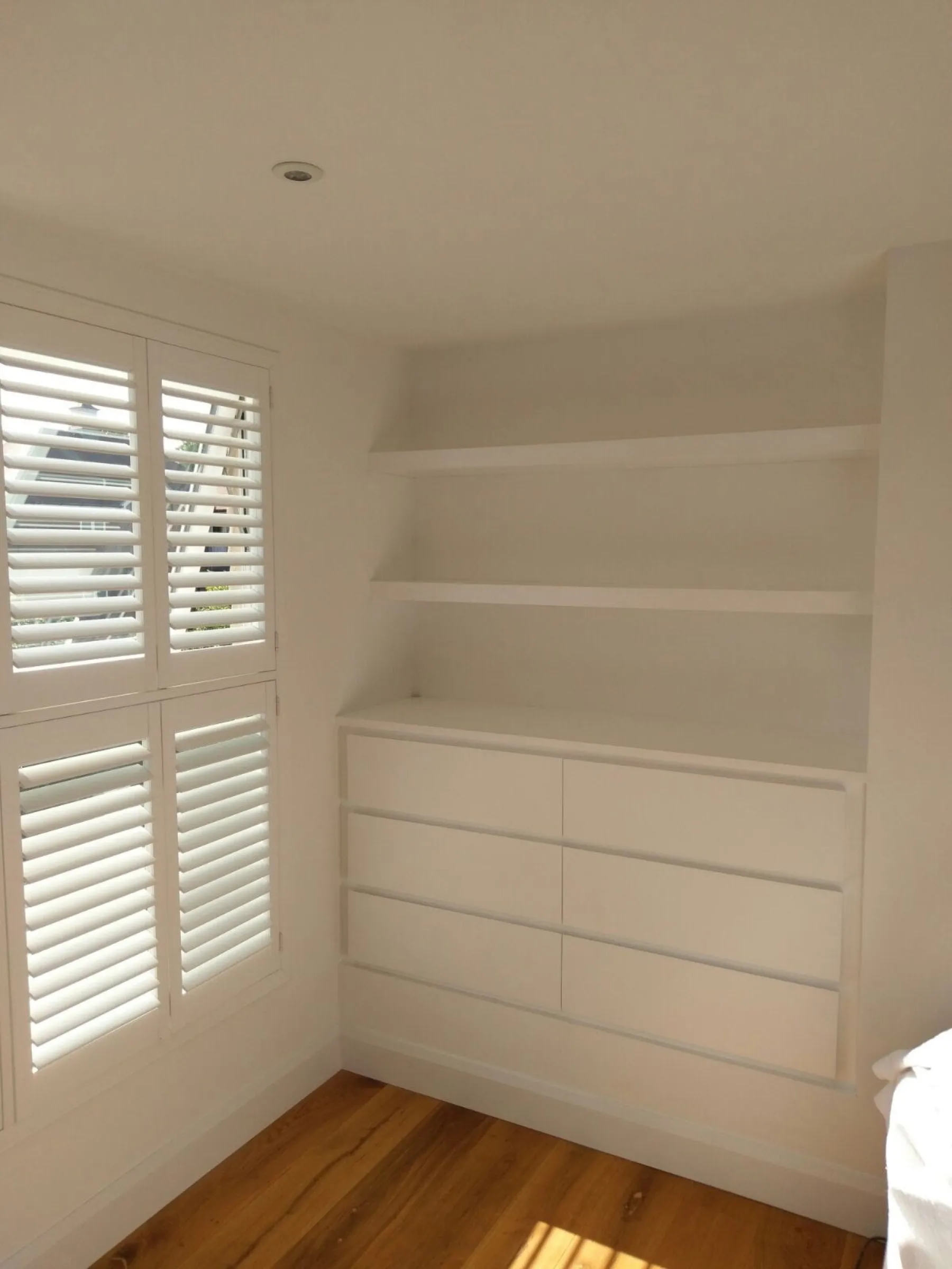 Finished fitted eaves drawer unit in white with floating shelves above and plantation shutters alongside