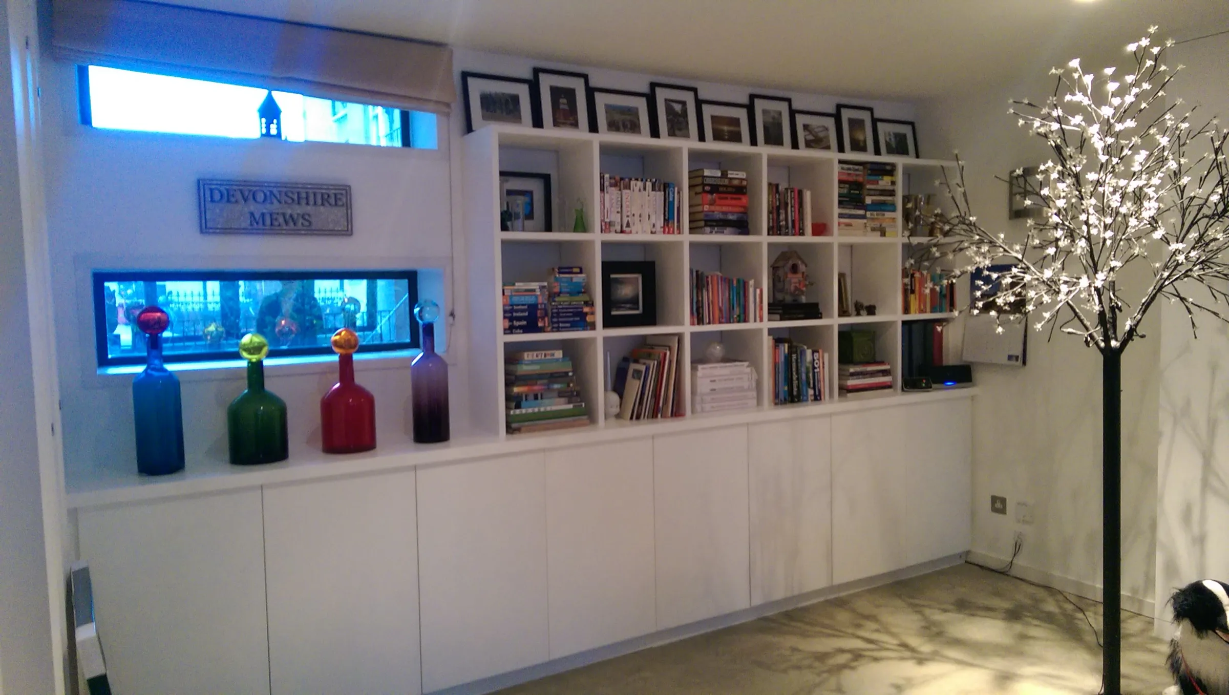 Contemporary wall of open box shelving above low handleless cupboards in a mews living room, styled with books and framed photographs