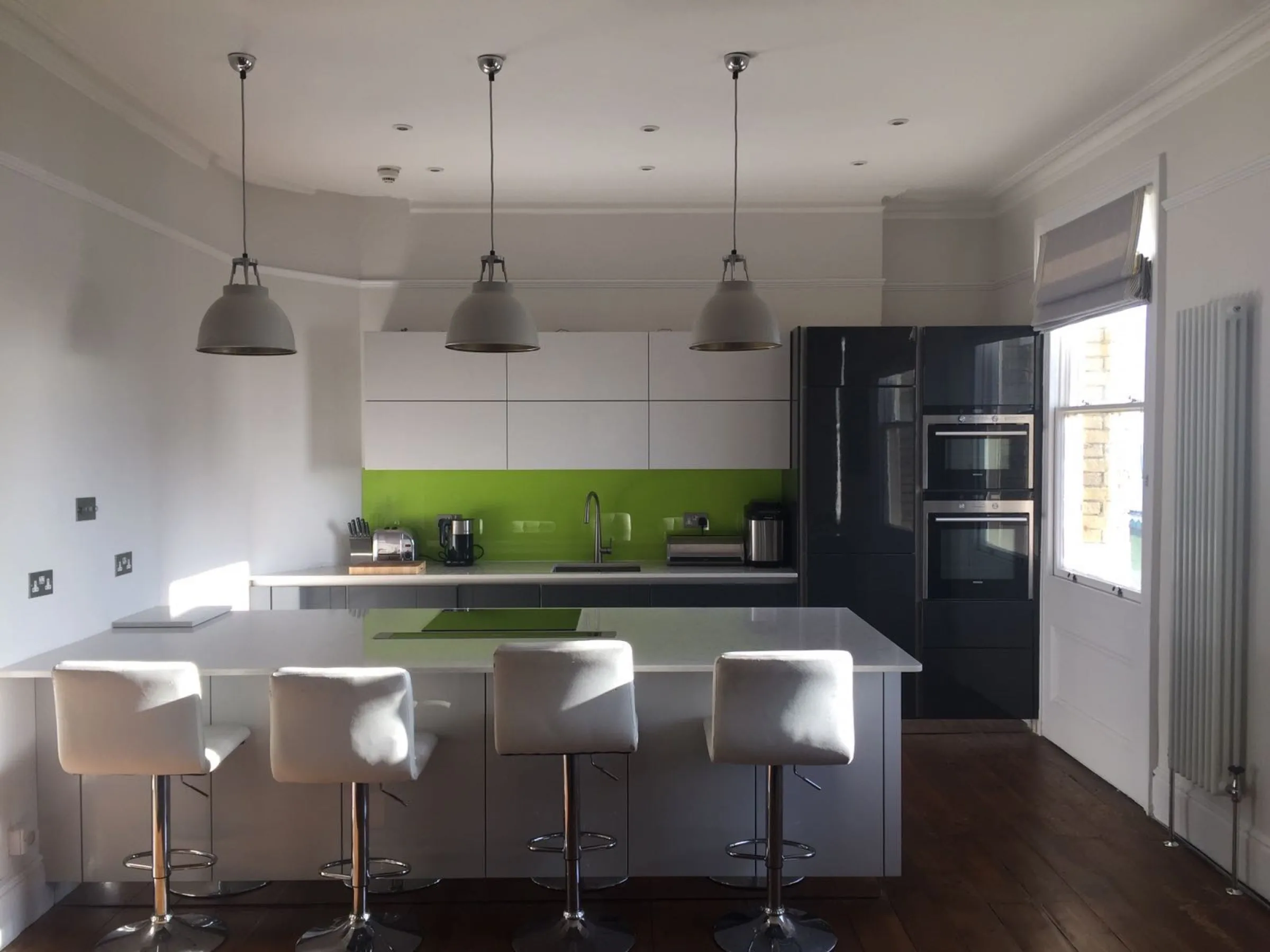 Open-plan kitchen in a period room with three industrial pendants over the quartz island, white bar stools and anthracite gloss tall units with integrated ovens