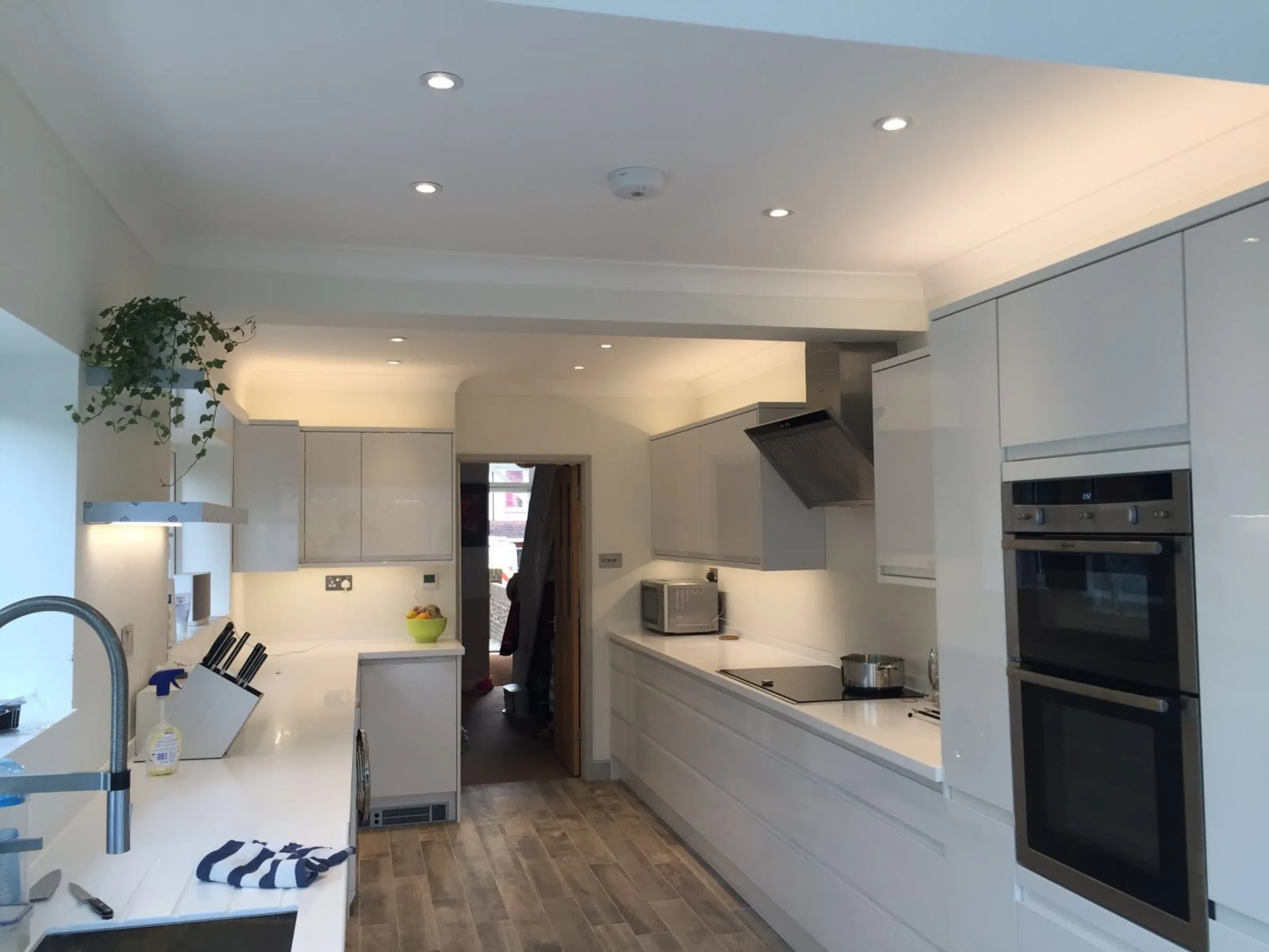 Galley kitchen with pale grey gloss handleless units, white worktops, eye-level double oven, stainless steel extractor hood and wood-effect floor, lit by under-cabinet lighting