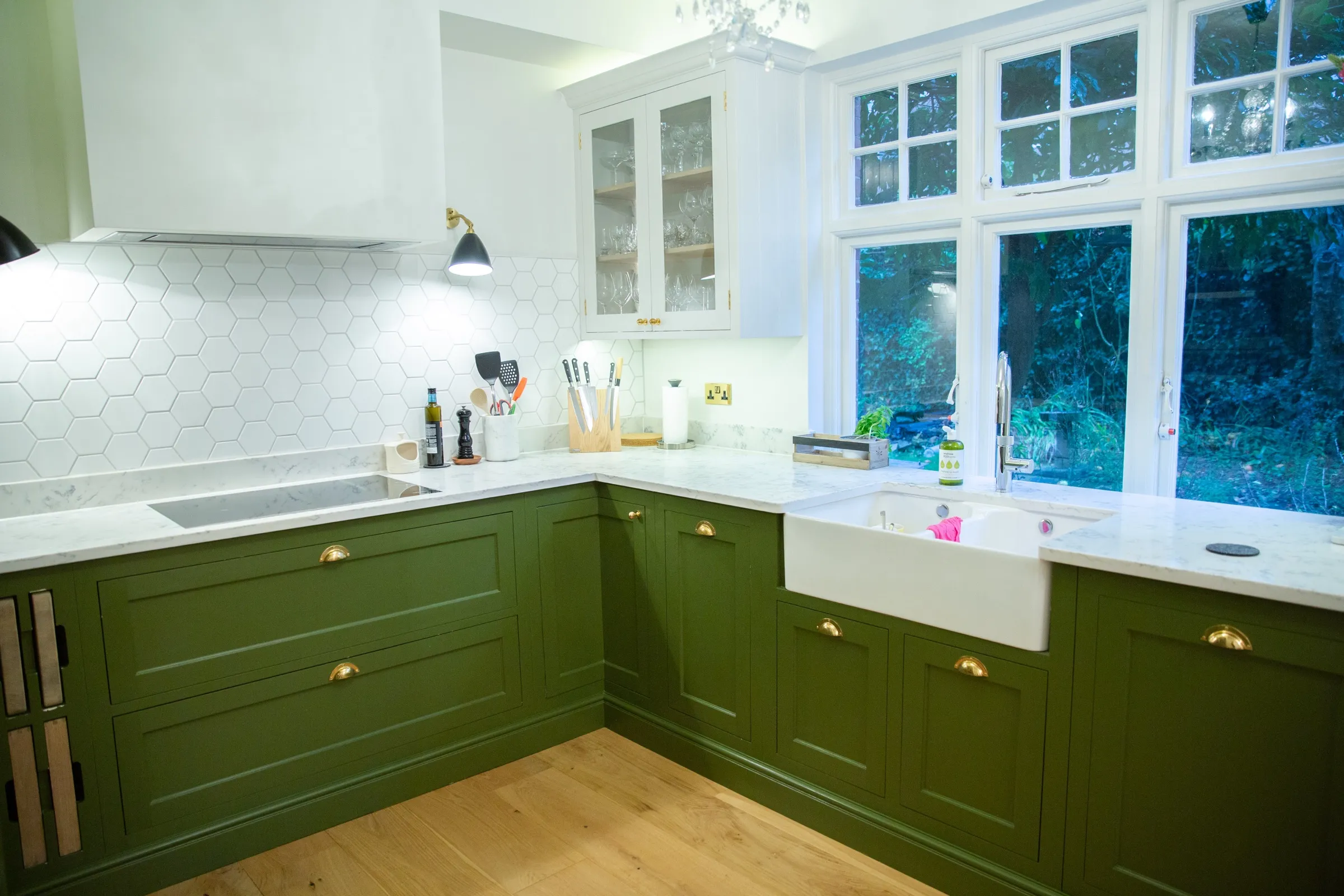 Corner of the finished kitchen with olive green base cabinets, brass cup handles, white hexagon splashback and a Belfast sink beneath the window