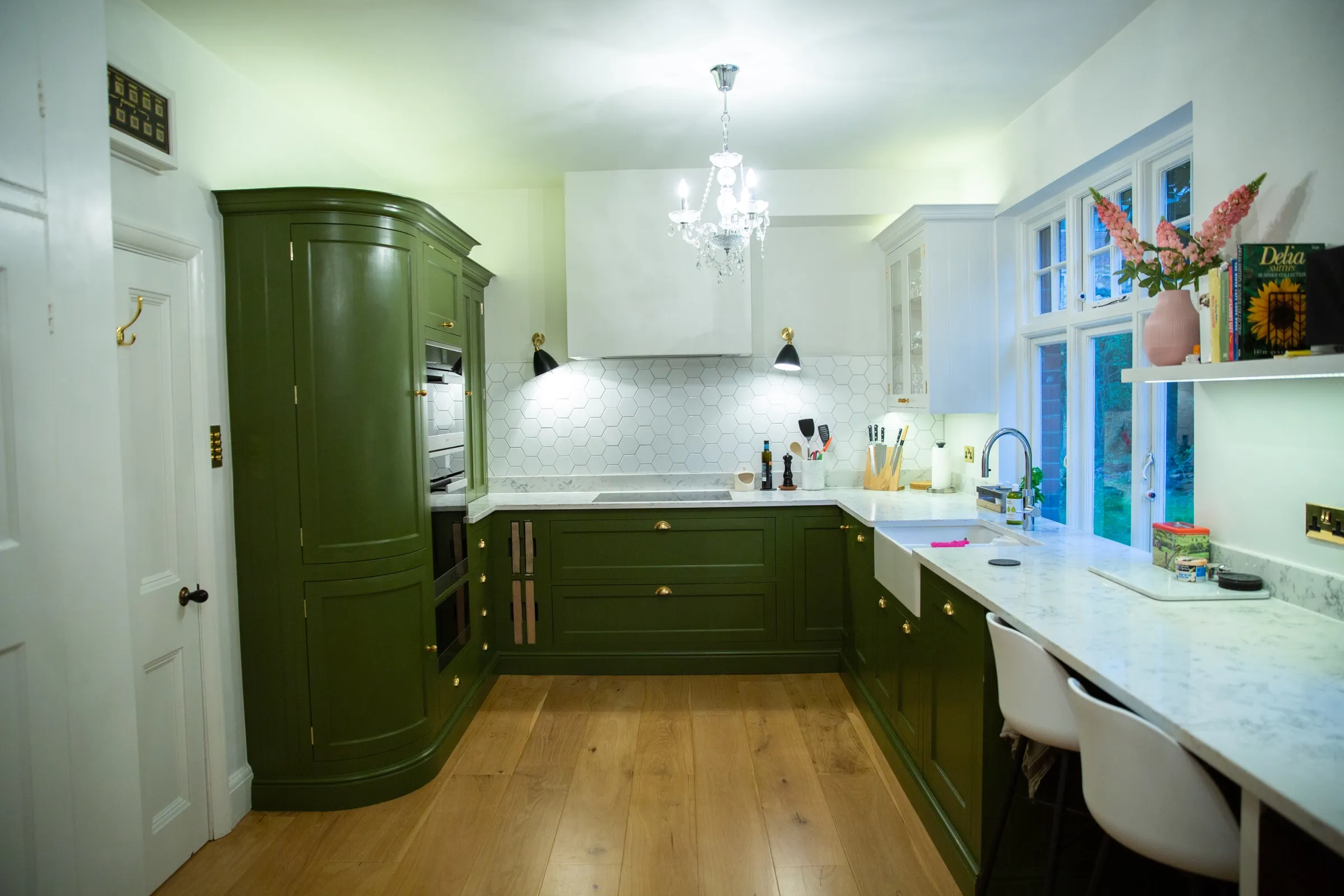 Full view of the hand-painted green kitchen with curved larder cabinet, glass chandelier, plastered extractor hood and oak flooring