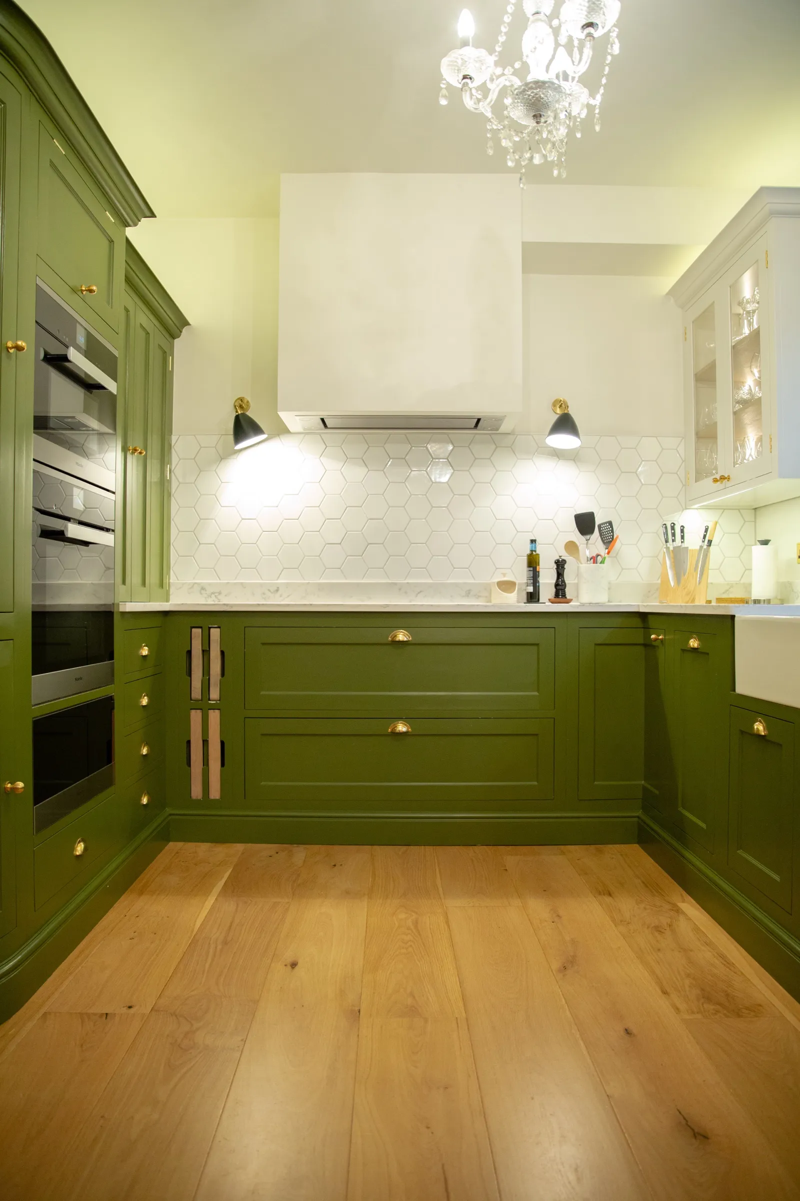 Tall green cabinets with integrated ovens beside the hob run, white extractor hood and hexagon-tiled splashback