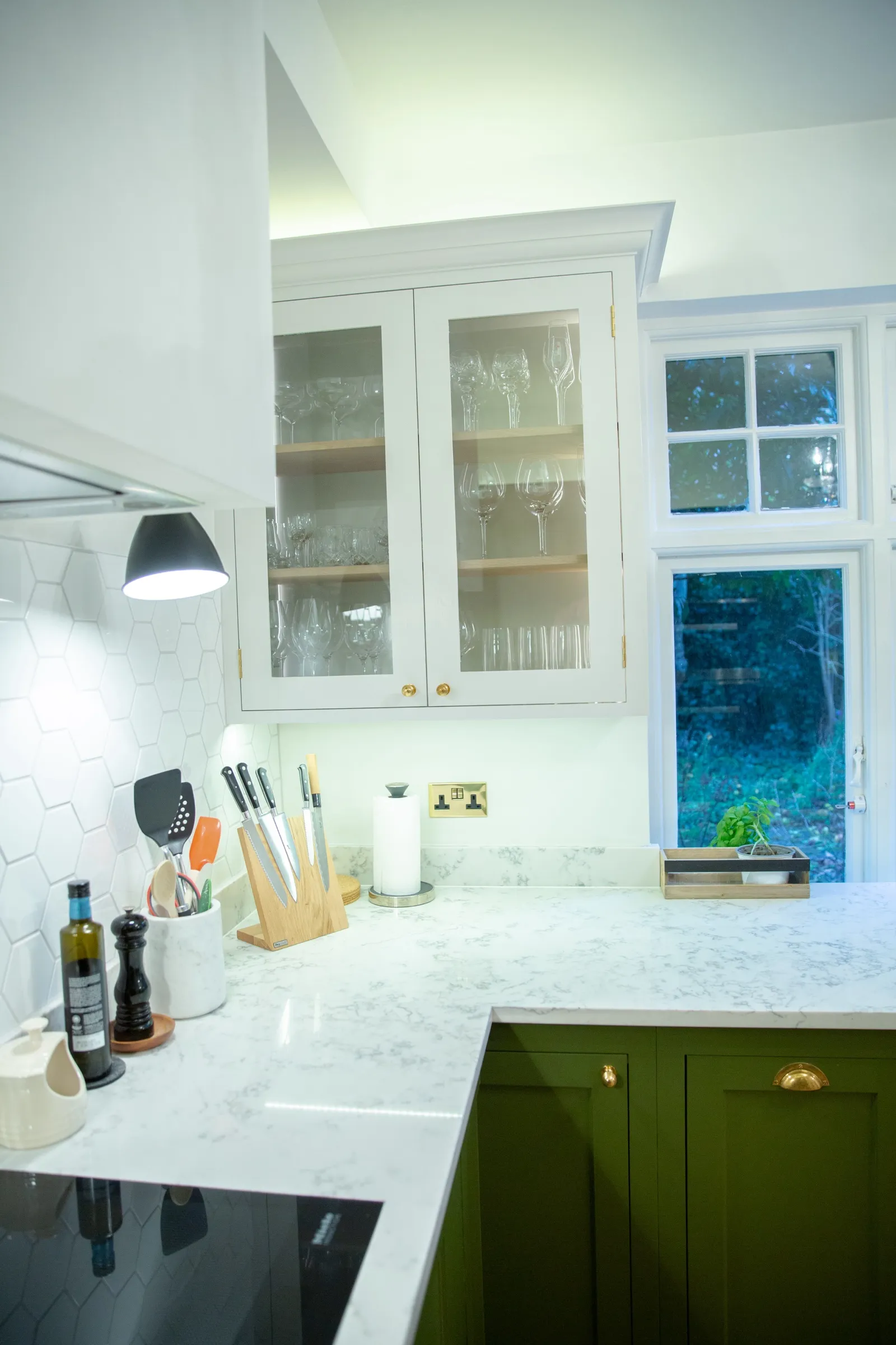 Glazed white wall cabinet with lit interior displaying glassware, above the quartz corner worktop and knife block