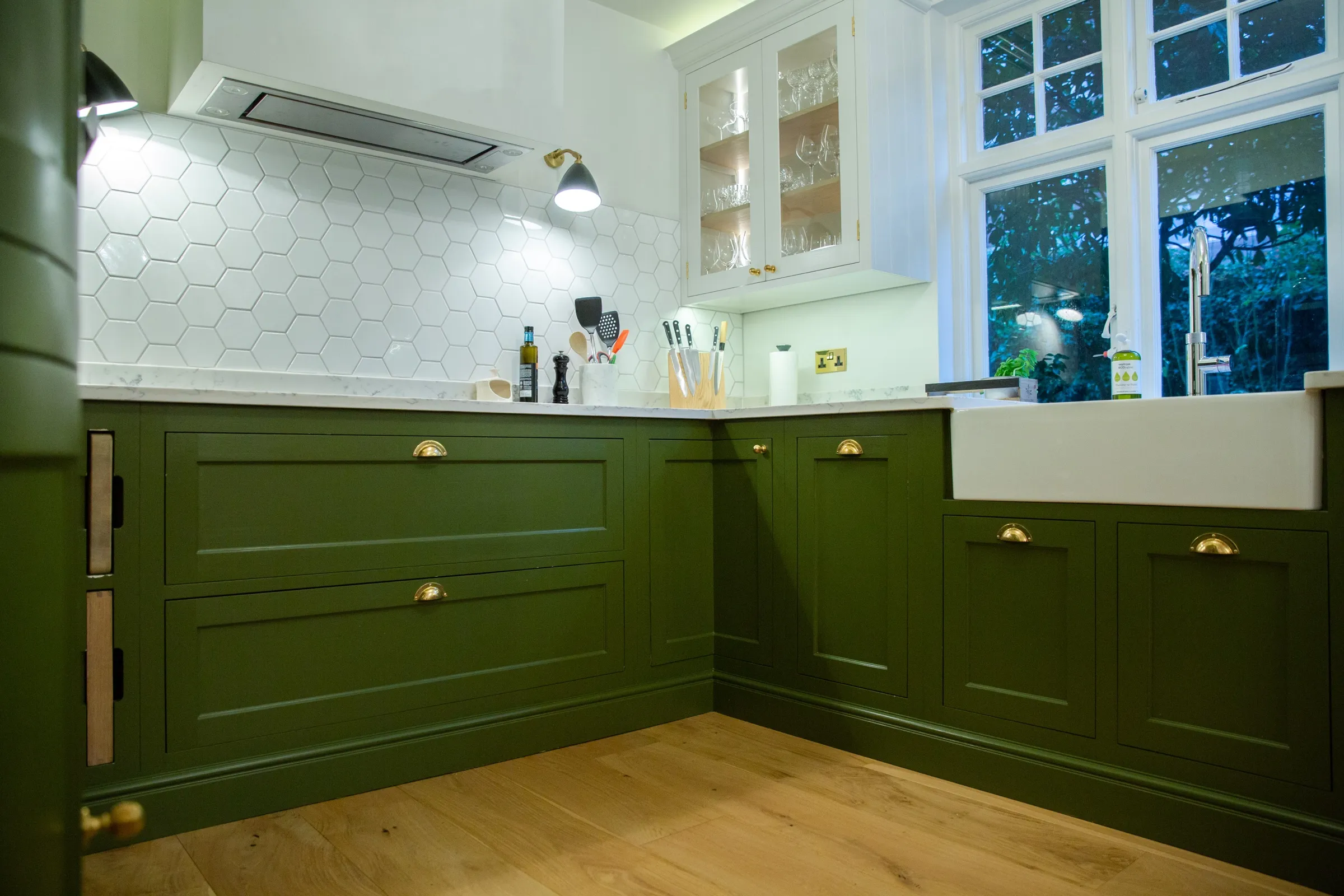 Long run of green base cabinets with Belfast sink, glazed wall cabinet and hexagon splashback, seen from the doorway