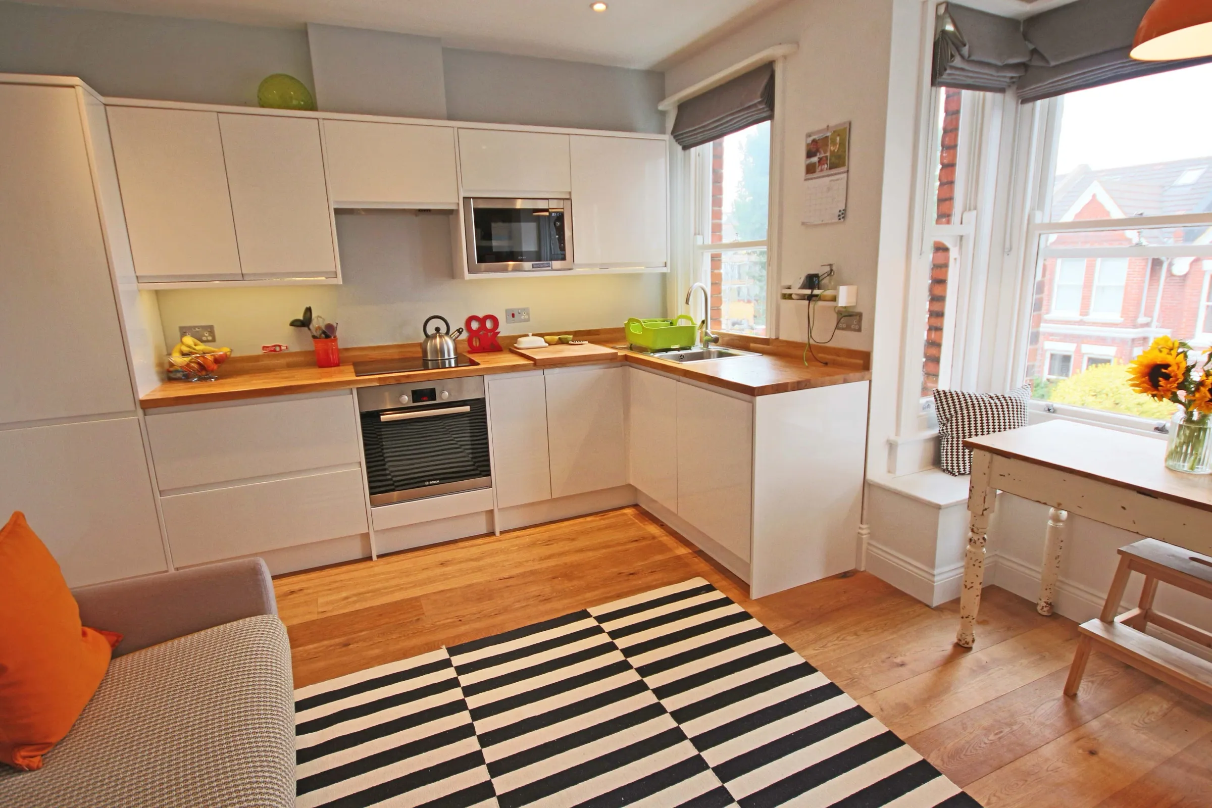 Wide view of an open-plan kitchen-diner with white handleless units, oak worktops and flooring, a striped rug, sofa and sash bay windows