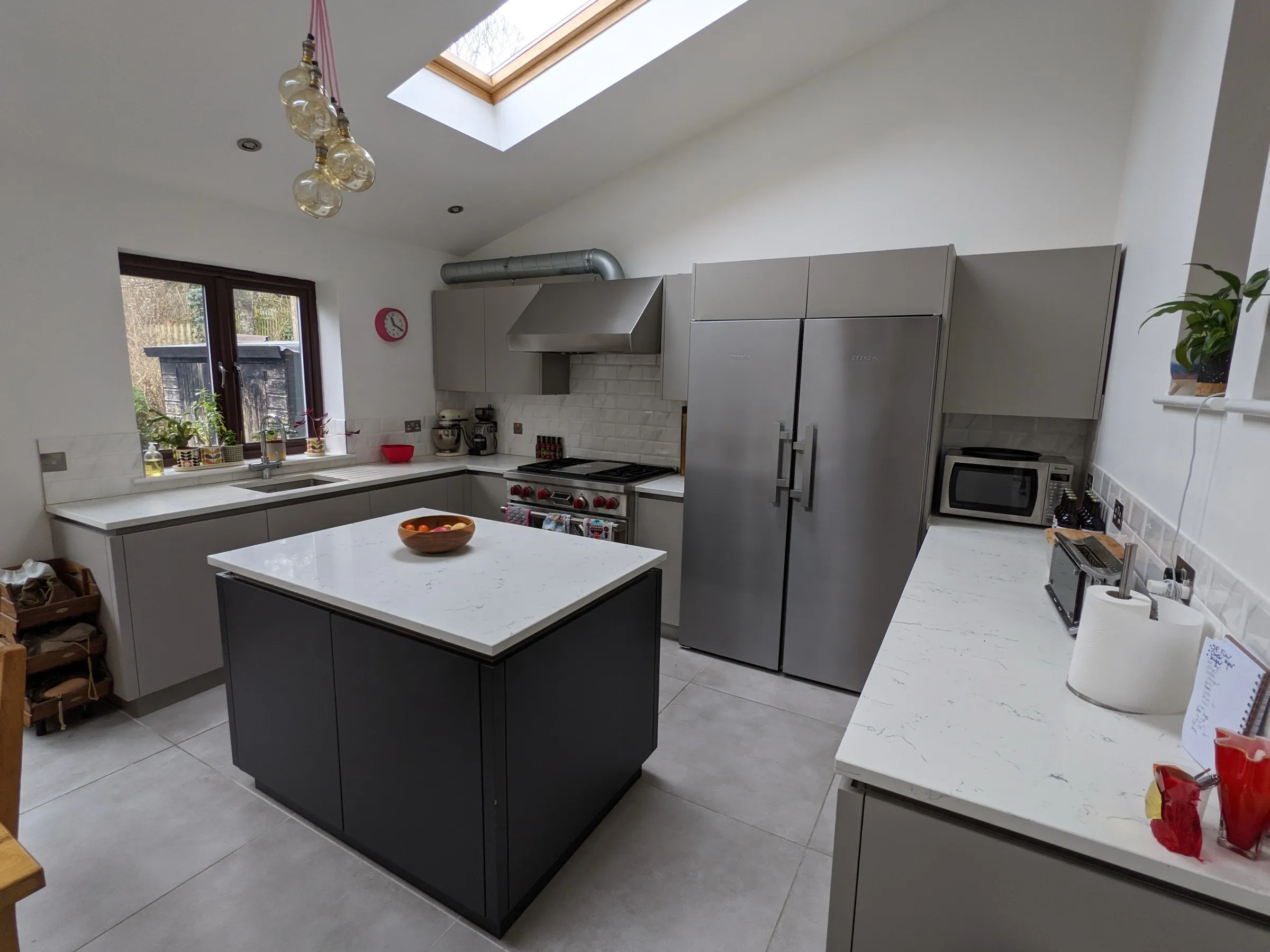 Light grey handleless kitchen with charcoal island, white quartz worktops and stainless steel range cooker beneath a vaulted skylight