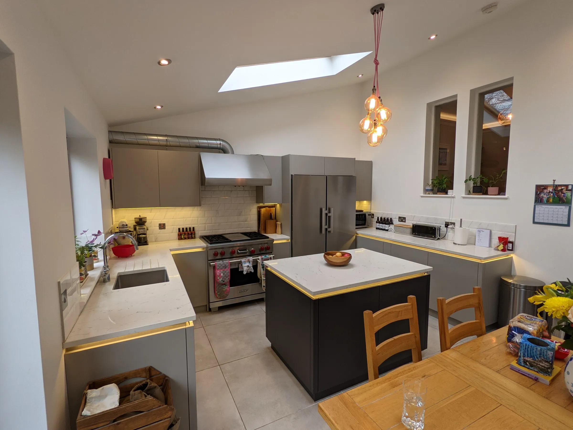 Wide view of the grey and charcoal island kitchen with pendant bulb cluster, skylight and oak dining table