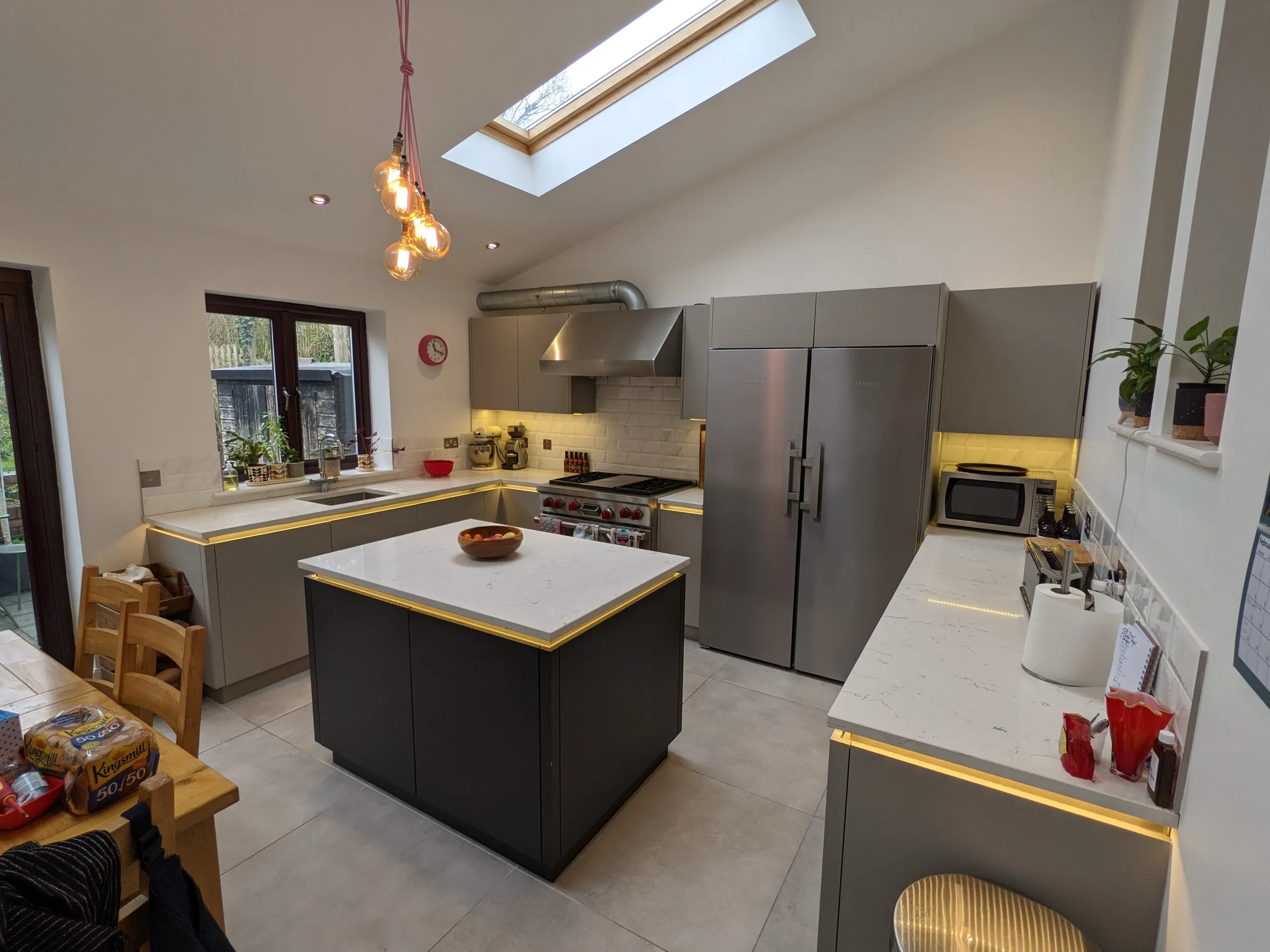 Olive green kitchen with white worktops and copper pendant lights during installation, beside a timber sash window