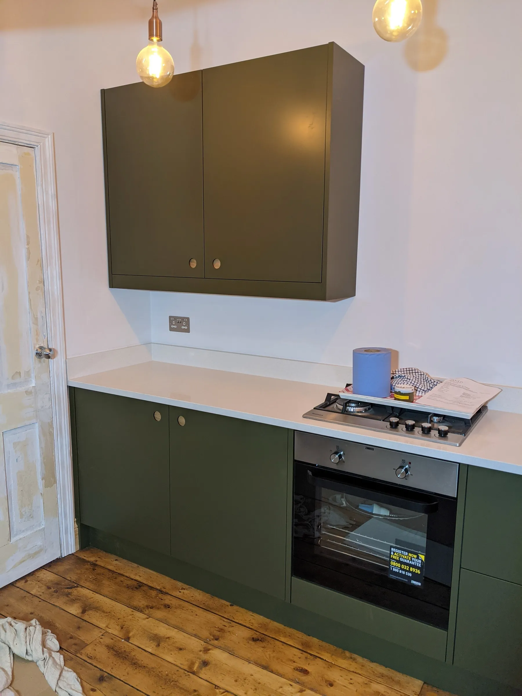 Full run of olive green handleless units and white worktops beneath three copper pendant bulbs, on stripped floorboards