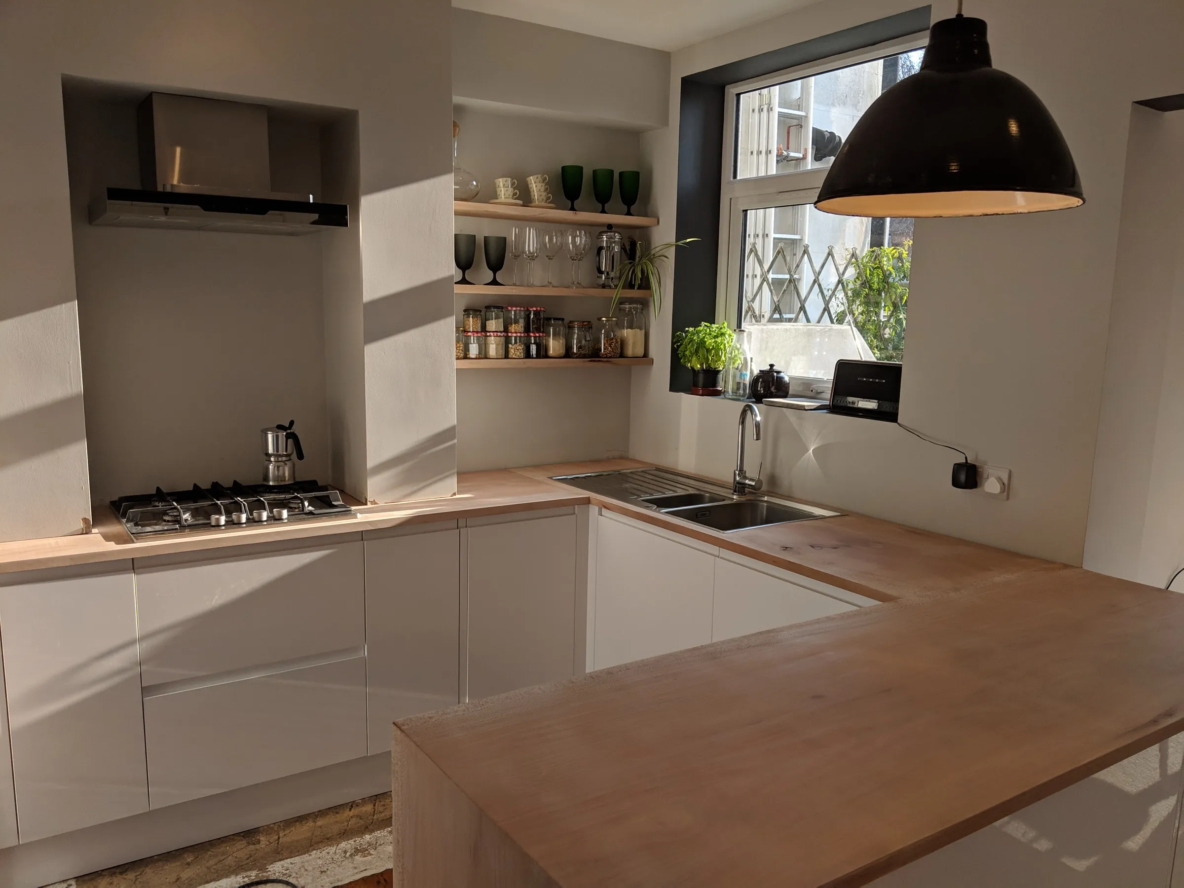 White handleless kitchen with pale timber worktops, open alcove shelving and breakfast bar beneath a black industrial pendant
