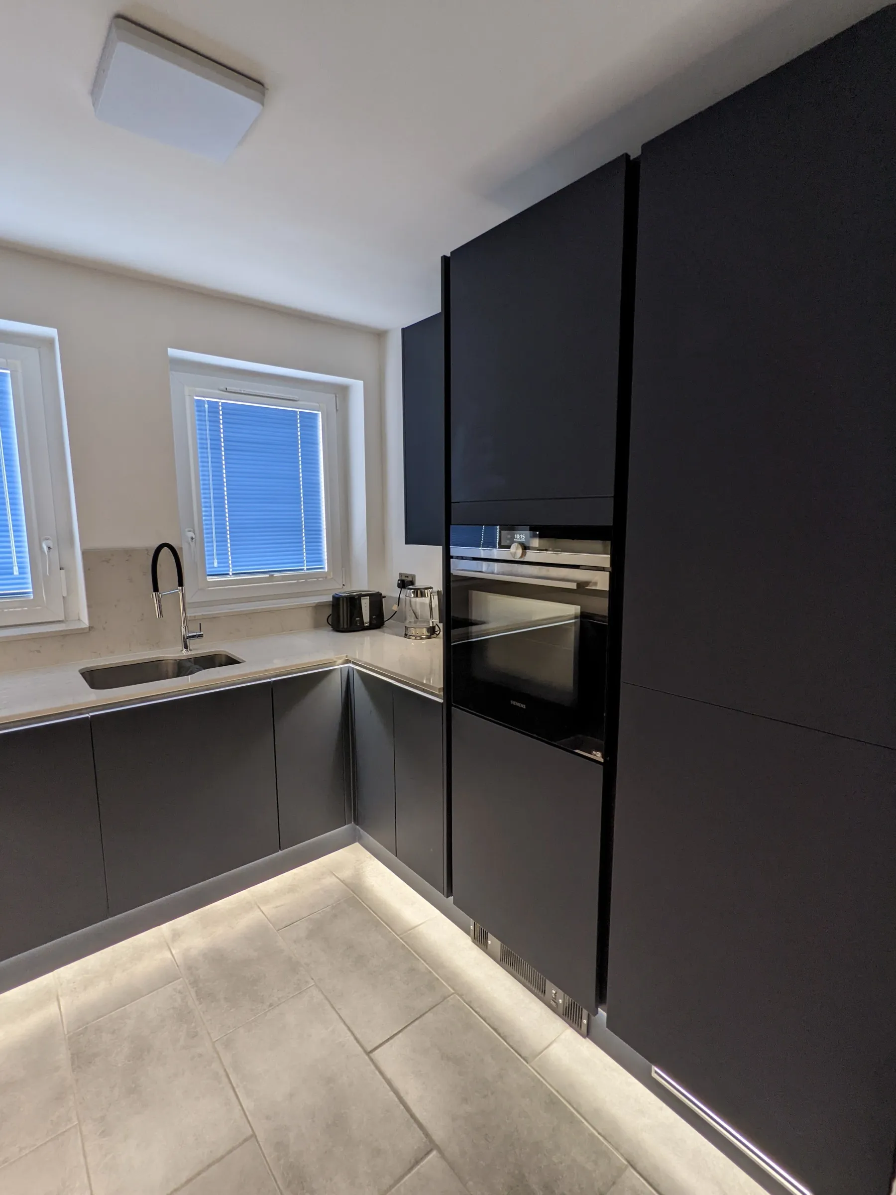 Matt black handleless kitchen with pale quartz worktop, integrated ovens and floor-washing LED plinth lighting