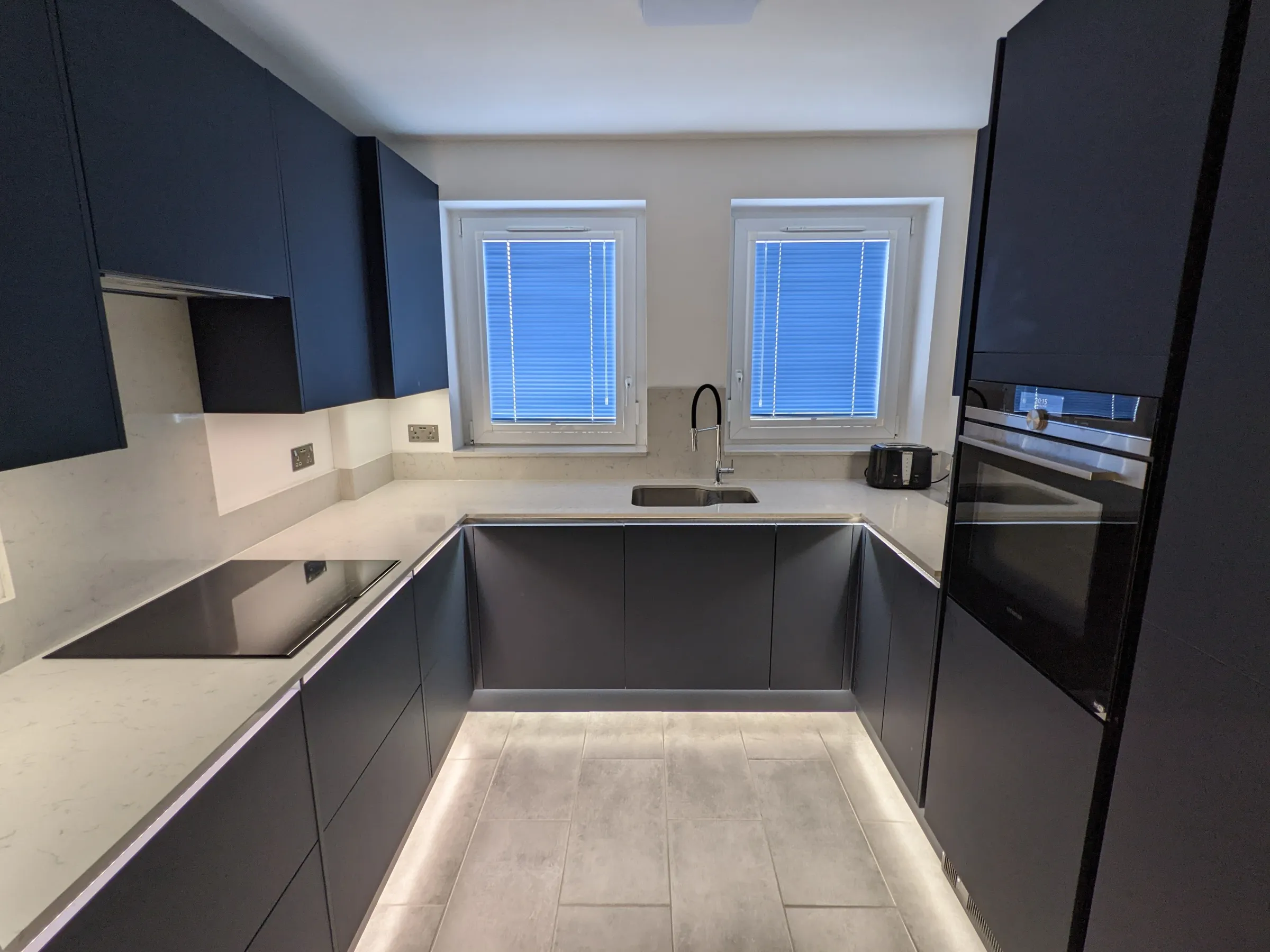 U-shaped matt black kitchen with white quartz worktops, twin windows and dramatic under-unit lighting