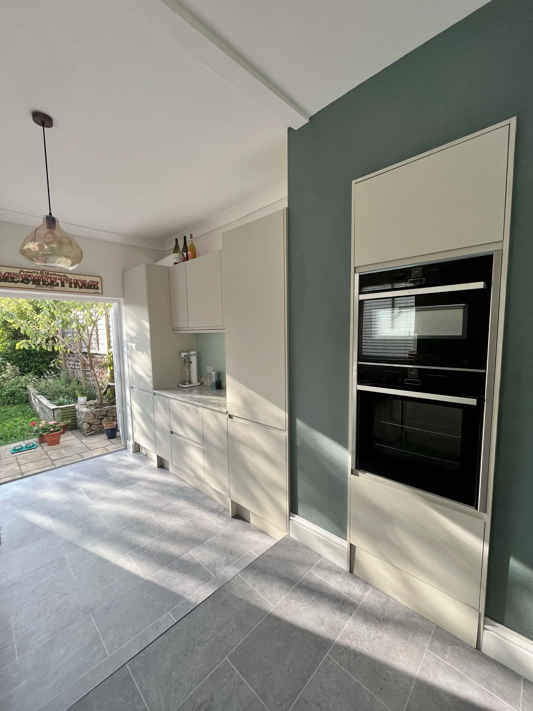 Cashmere kitchen with larder run, built-in ovens on a teal wall and doors opening to the garden