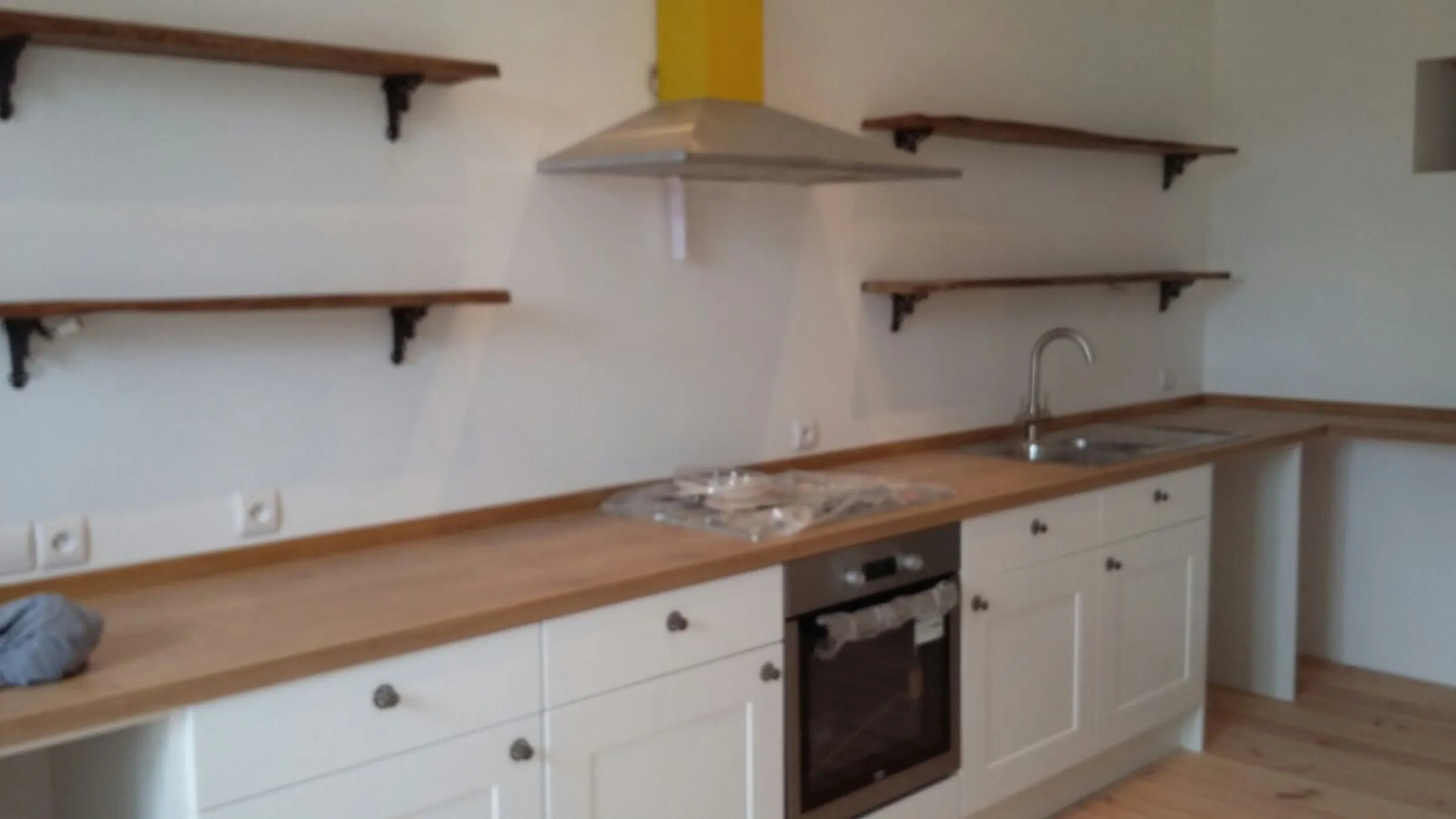 Long run of cream Shaker units and oak worktops mid-installation, with waney-edge timber shelves on cast iron brackets and a stainless chimney hood