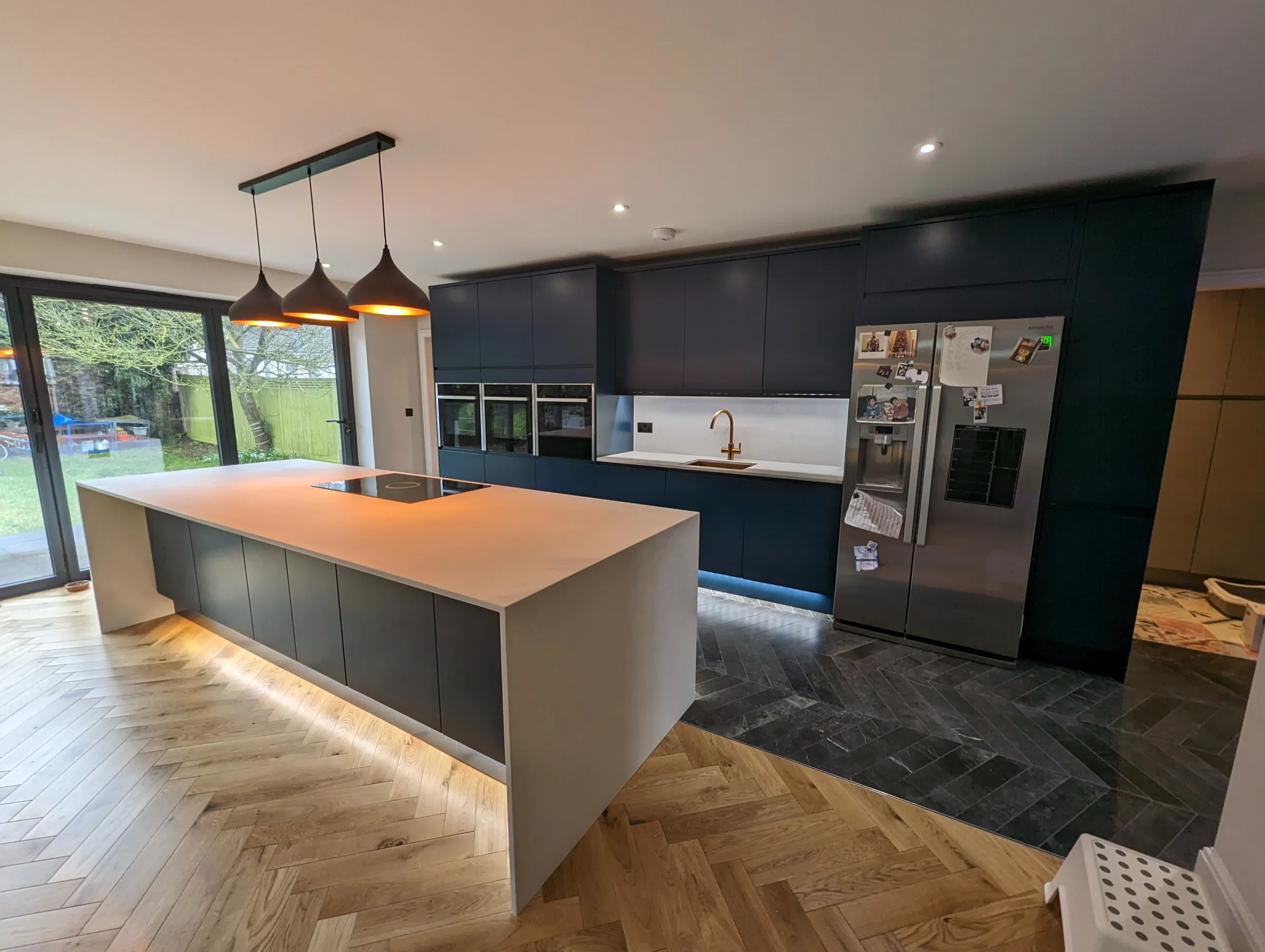 Open-plan navy kitchen with white waterfall island, bifold doors and herringbone oak floor
