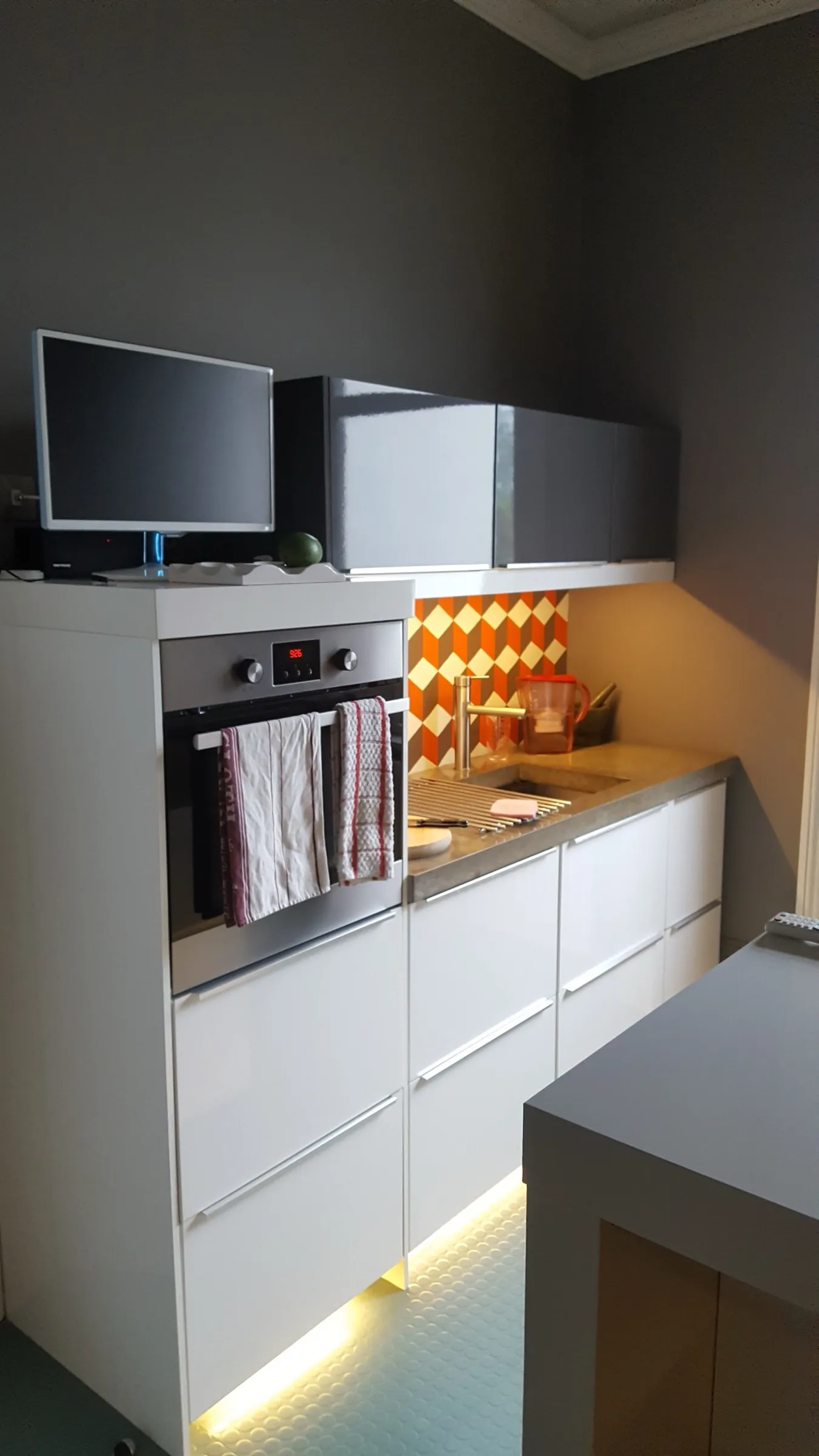 White gloss drawer units and concrete-style worktop with a retro orange geometric splashback, grey gloss wall cabinets and warm under-unit lighting