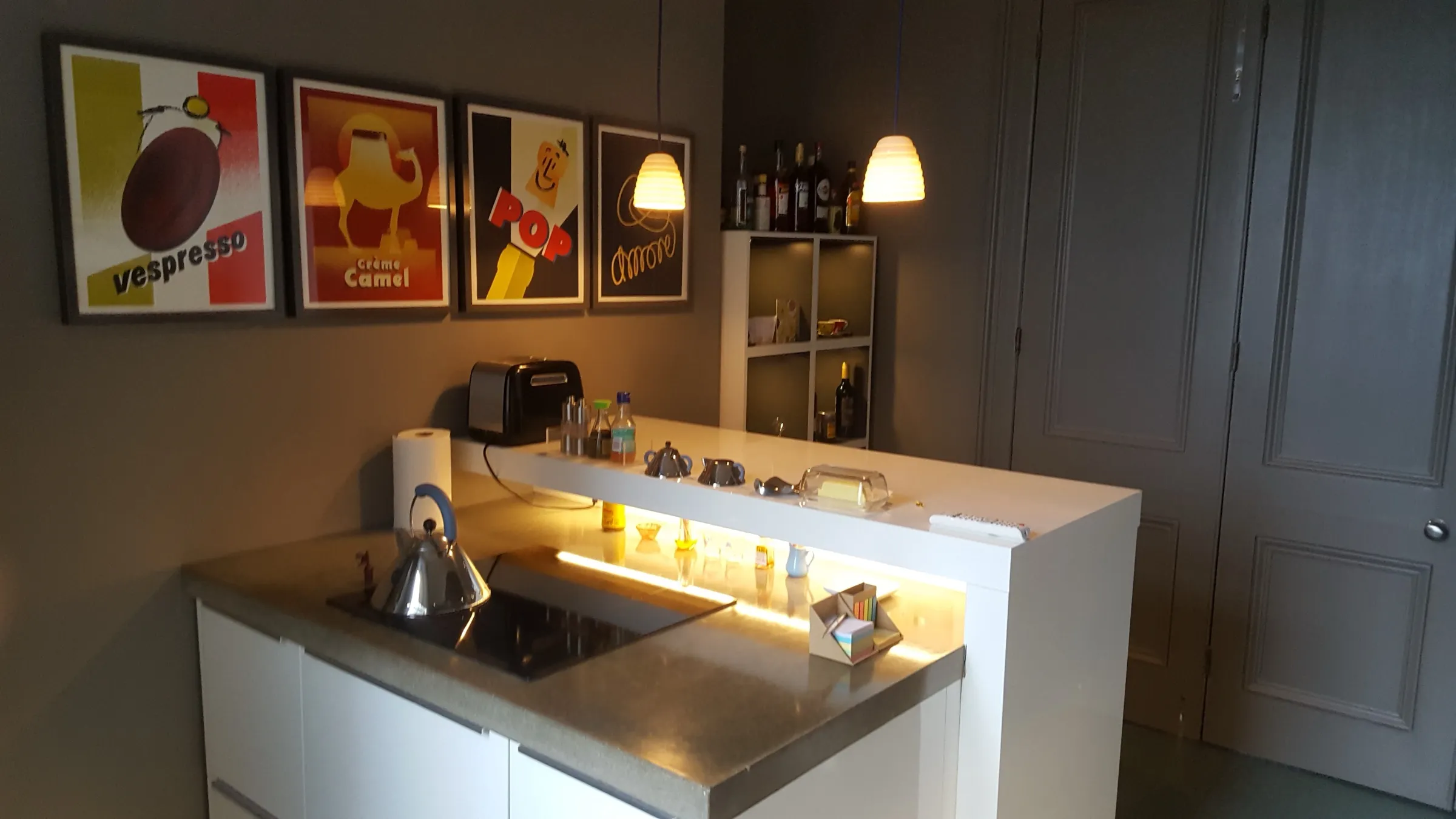 Raised white breakfast bar with concealed LED lighting and beehive pendant lights, beside framed retro coffee prints on a dark grey wall
