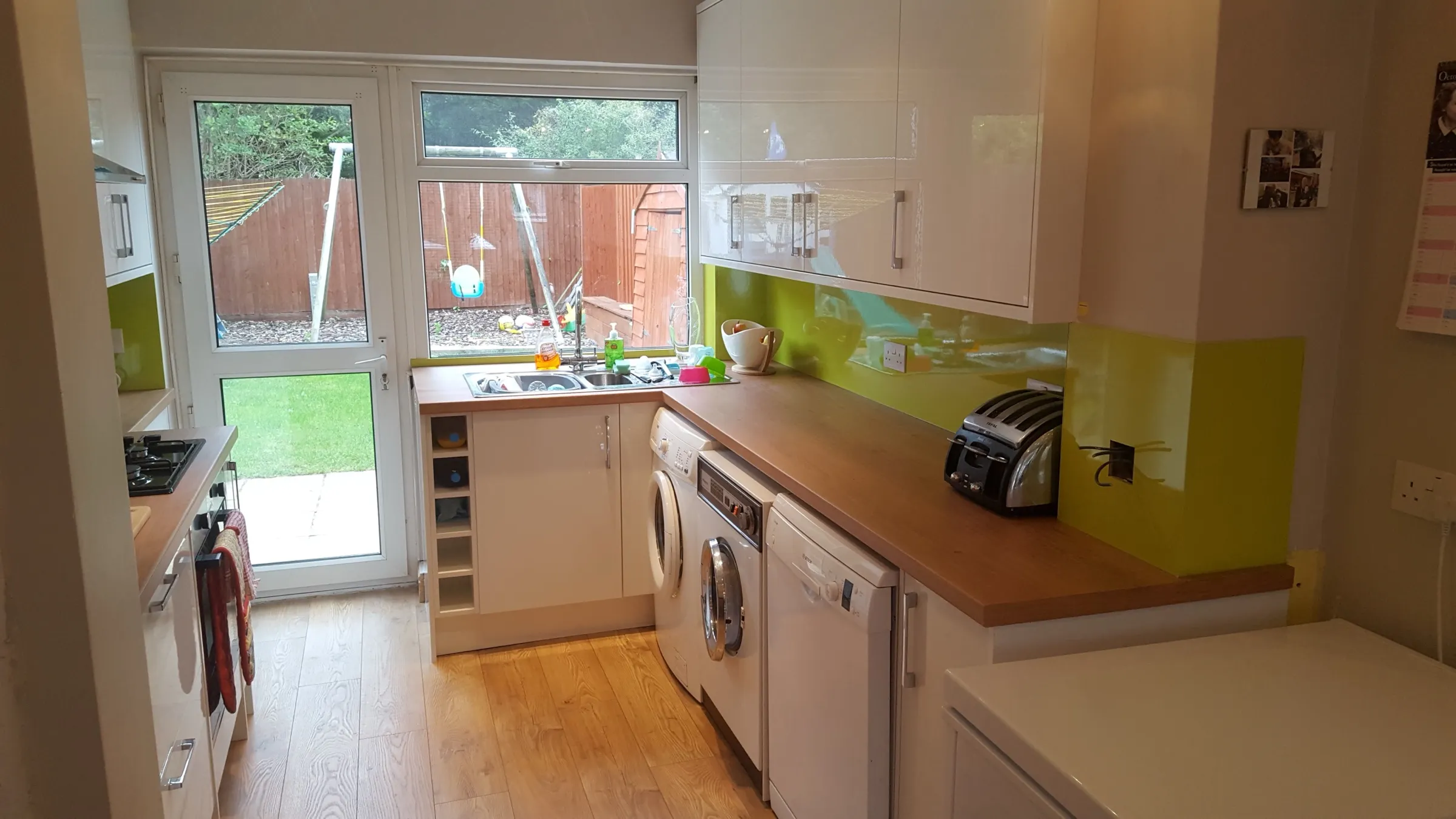 White gloss kitchen with lime green glass splashbacks, wood-effect worktops, wine rack storage and sink beneath a window overlooking the garden