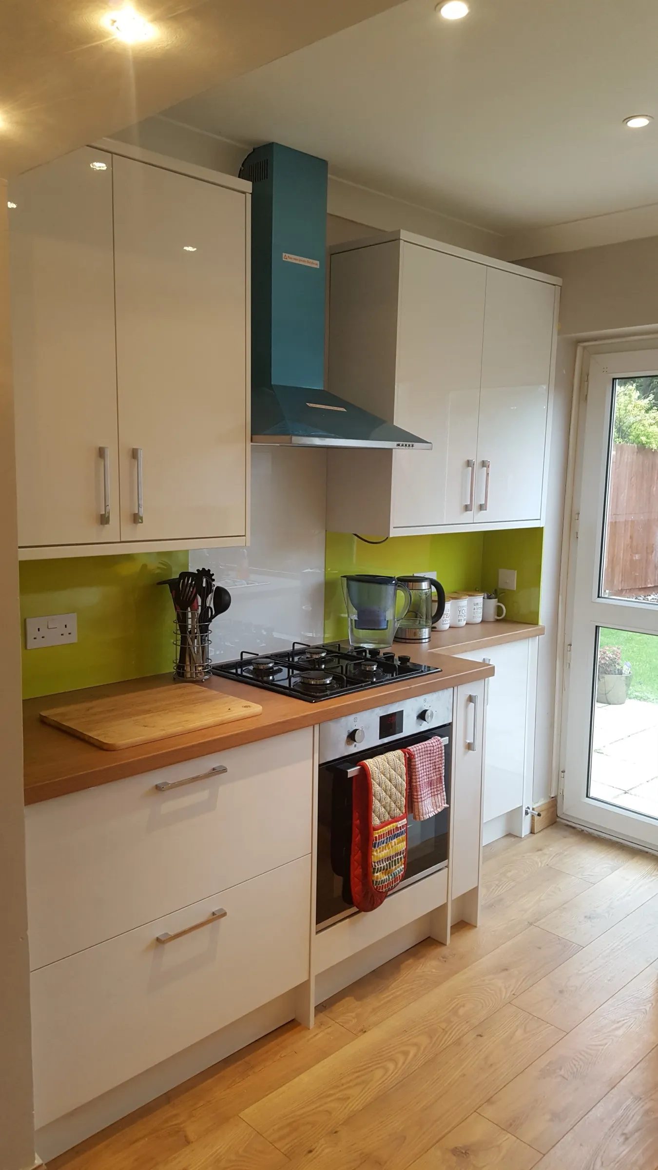 Cooker run with black gas hob, built-in oven, teal chimney extractor hood and lime green glass splashback against white gloss units