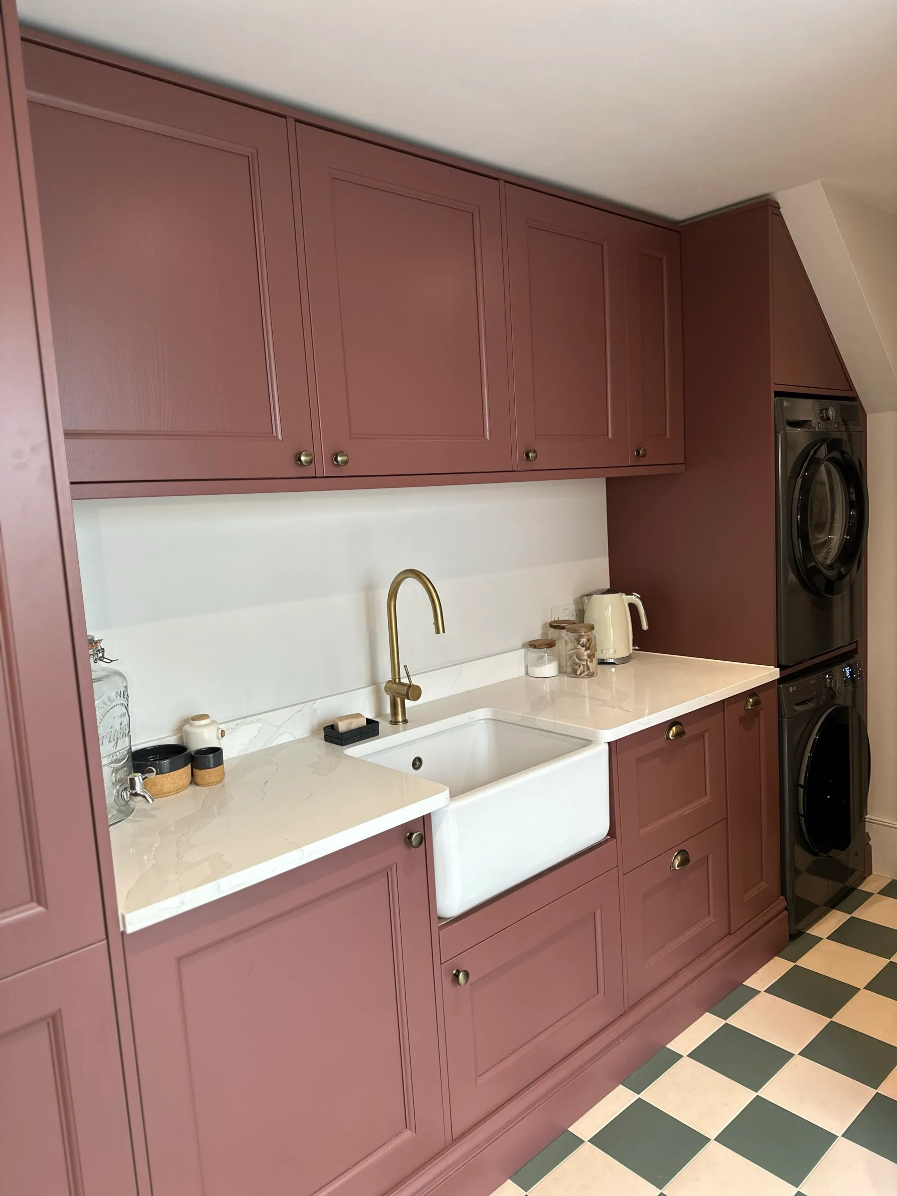 Angled view along the maroon utility cabinetry towards the Belfast sink, brass tap and stacked laundry appliances