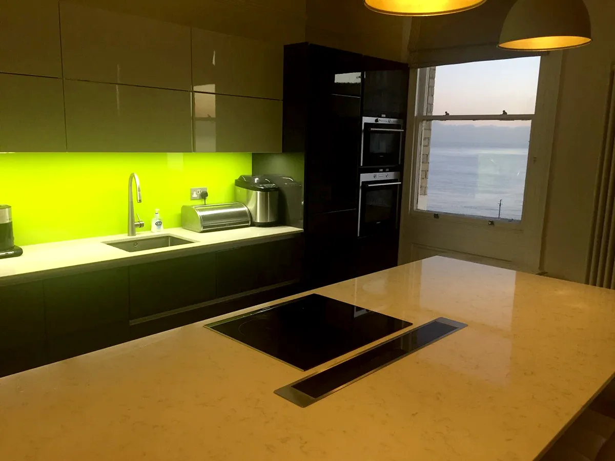 Quartz kitchen island with induction hob and downdraft extractor in a seafront apartment, with backlit green splashback and sea view from the sash window