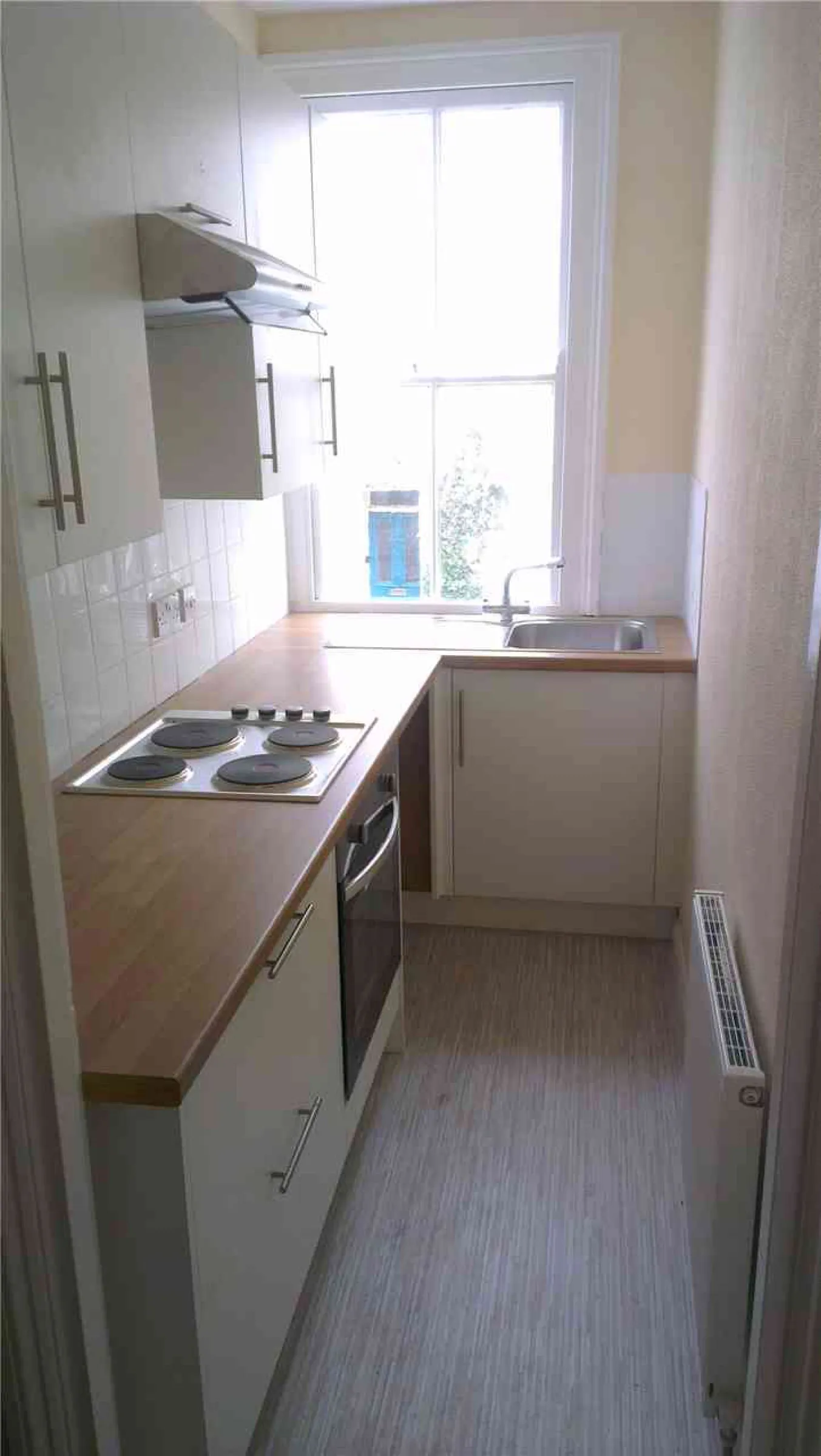 Compact galley kitchen with cream units, wood-effect worktops, four-ring electric hob beneath a stainless steel canopy hood, and sink under a sash window
