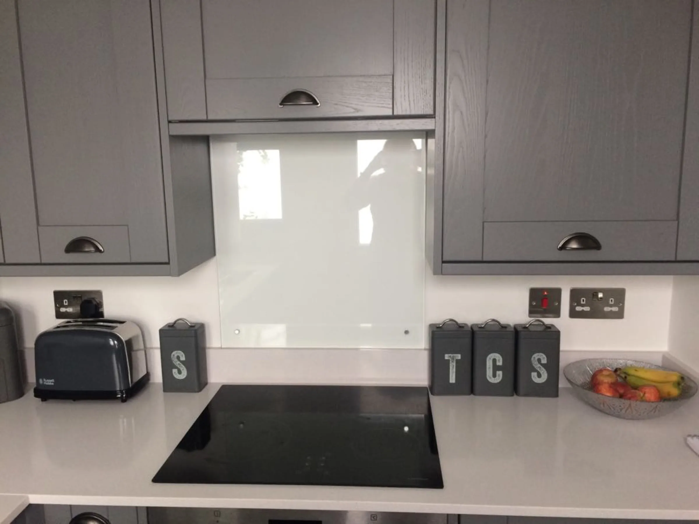 Grey Shaker wall cabinets with pewter cup handles above a black hob and white glass splashback