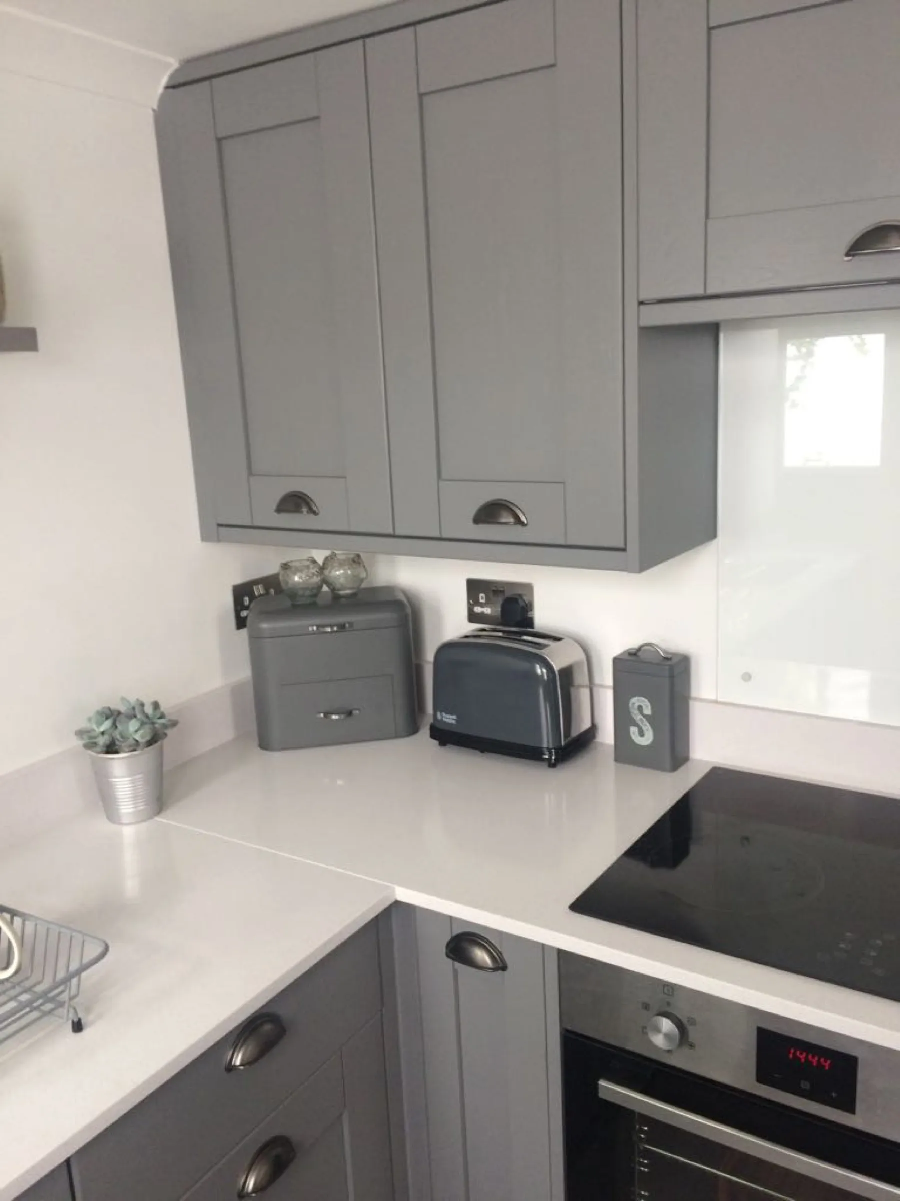 Corner of a grey Shaker kitchen with built-under oven, black hob and matching grey bread bin and toaster
