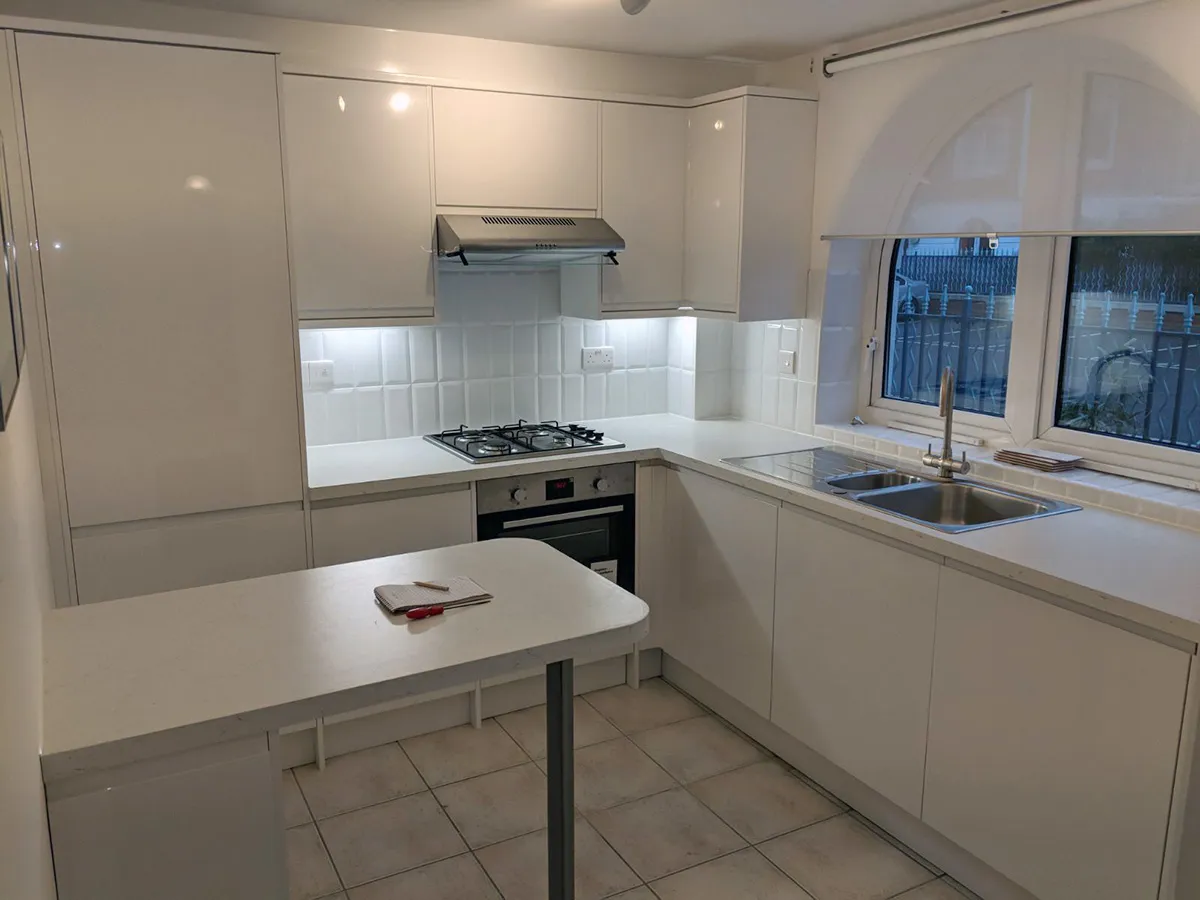 Compact L-shaped white gloss kitchen with metro-tiled splashback, stainless steel hob and hood, and a small breakfast table